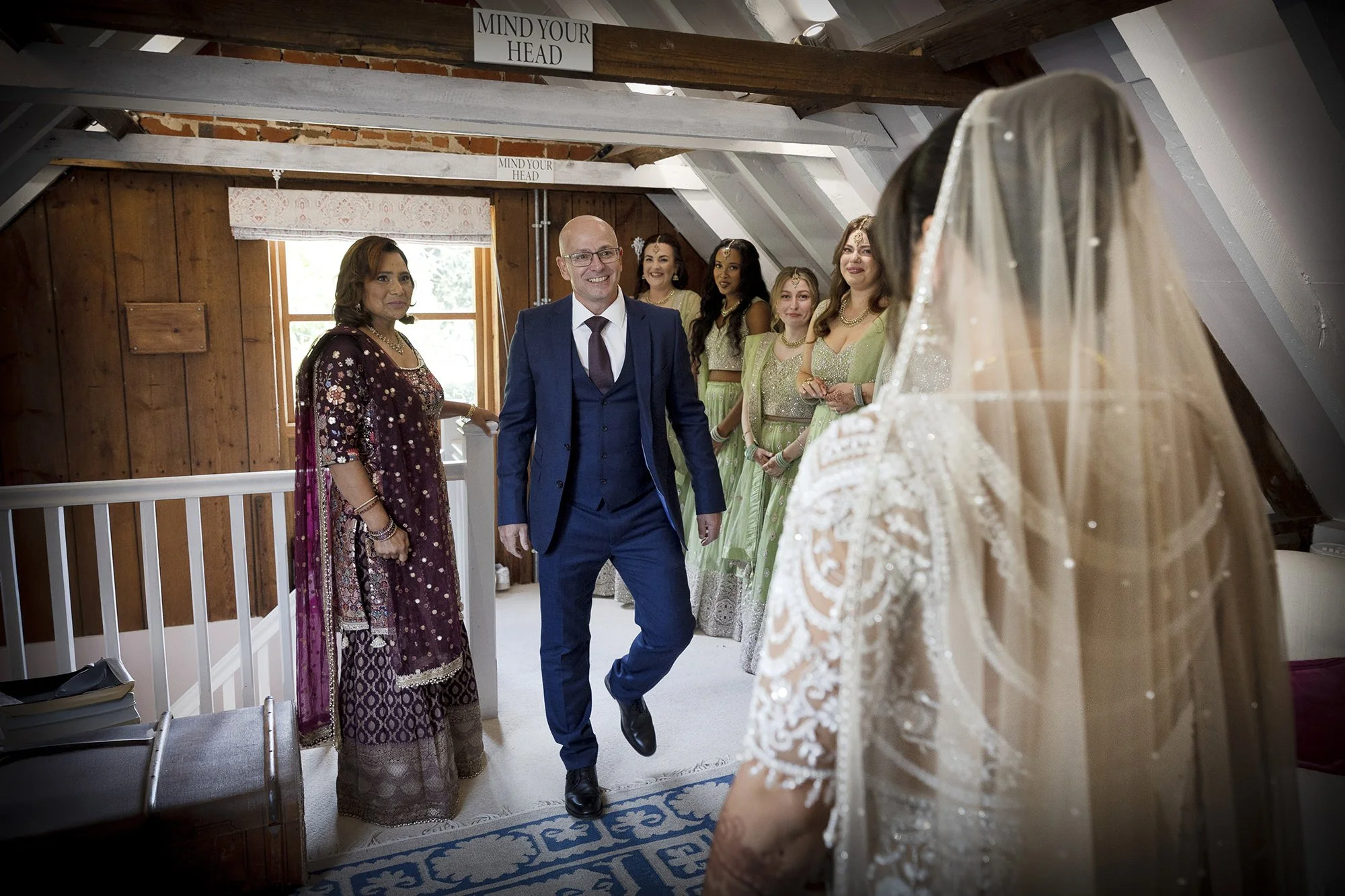 A bride in a white lace wedding dress and veil stands facing a smiling man in a blue suit as he enters a room. Four women in green traditional dresses watch, one woman in a purple and gold traditional dress stands nearby. The room has wooden walls, a