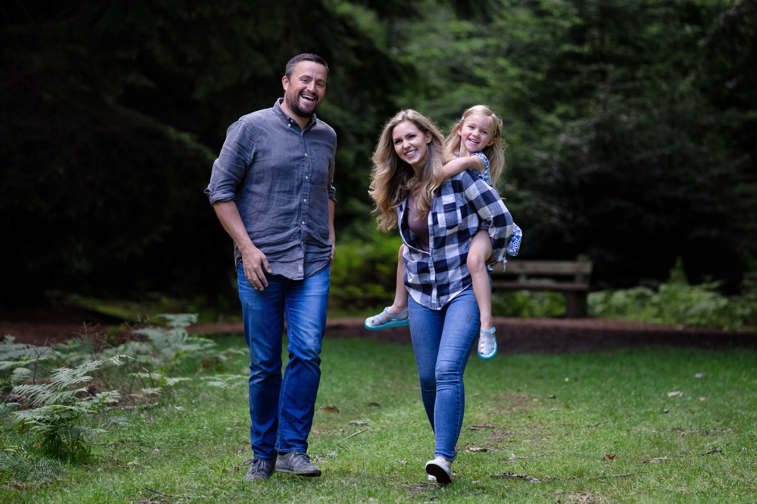 A family of three, including a man, woman, and young girl, smiling and walking in a park with greenery and trees in the background.