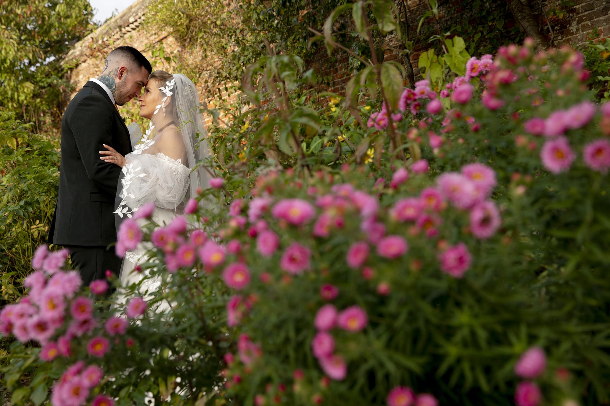 Bride and groom in wedding attire standing close together, with their foreheads touching, surrounded by pink flowers and greenery.