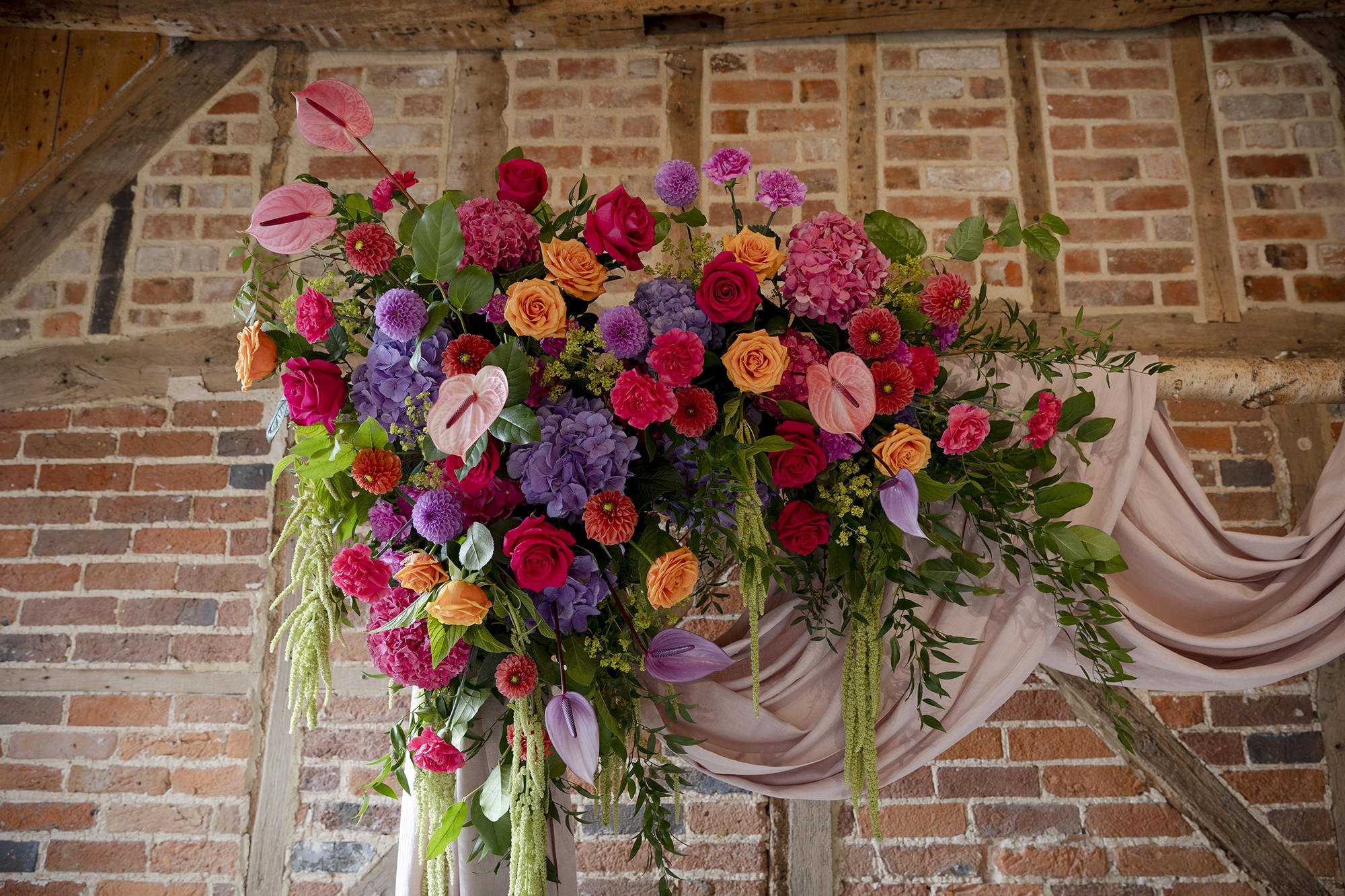Large floral arrangement with pink, purple, orange, and red flowers, draped with pink fabric, against a brick and wood ceiling background.