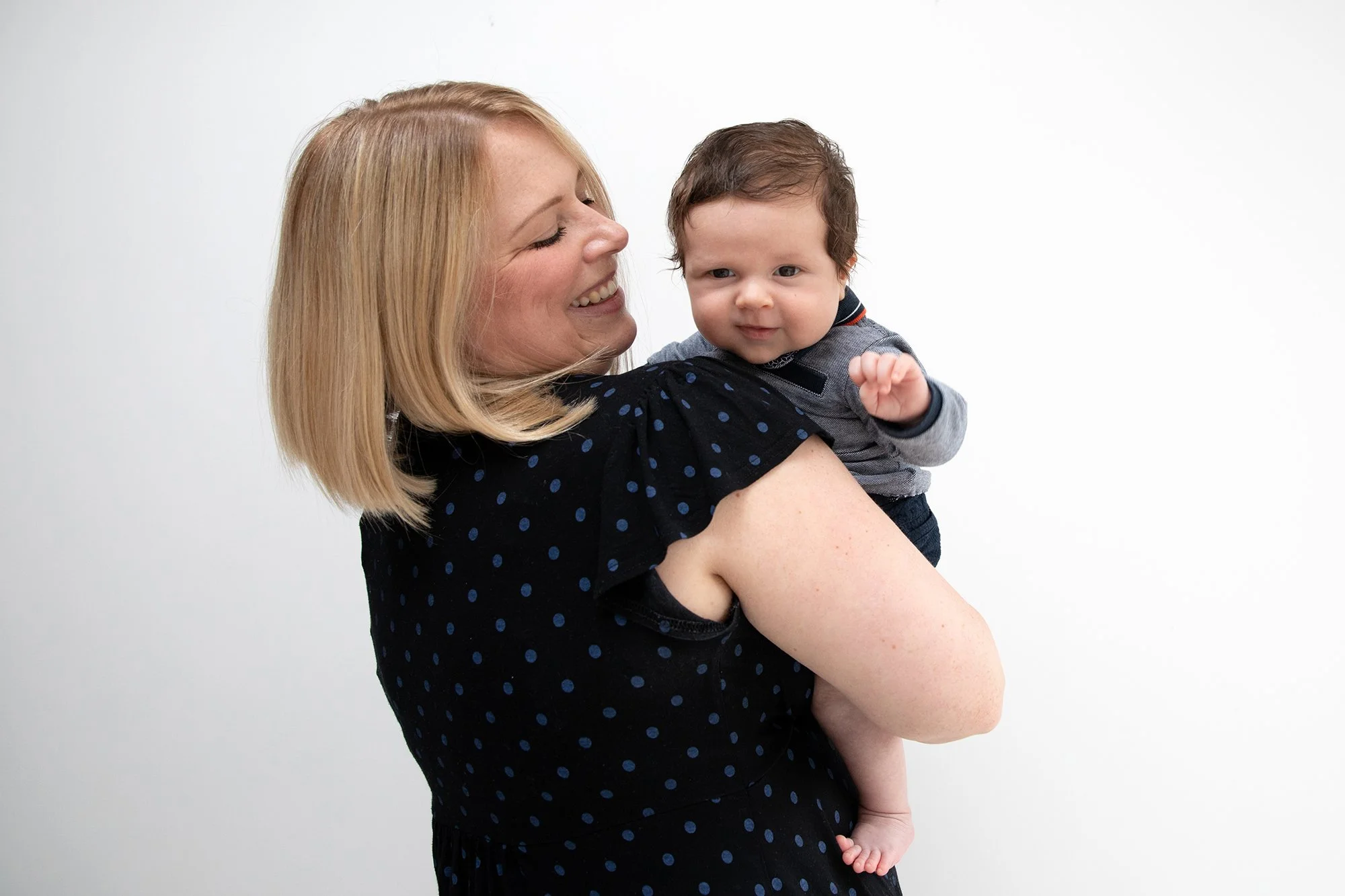 A woman with blonde hair holding a young child with dark hair, both smiling against a plain white background.