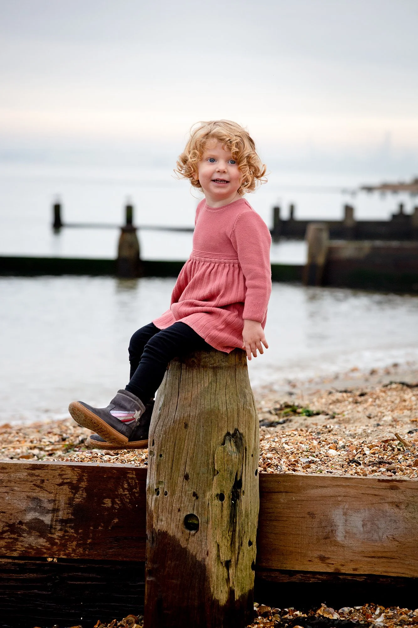 A young girl with curly blonde hair wearing a pink dress, black leggings, and gray shoes sitting on a wooden post by the water at the beach during sunset.