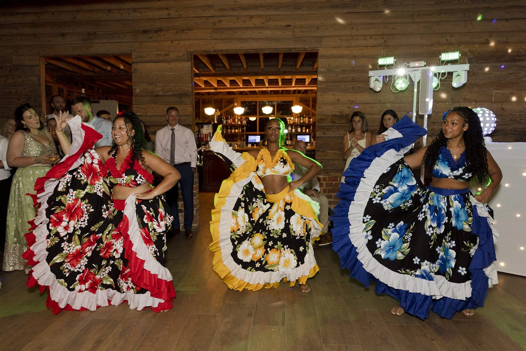 Three women in colorful, floral-patterned skirts performing a traditional dance at a social event, with a lively crowd watching in the background inside a rustic wooden venue.