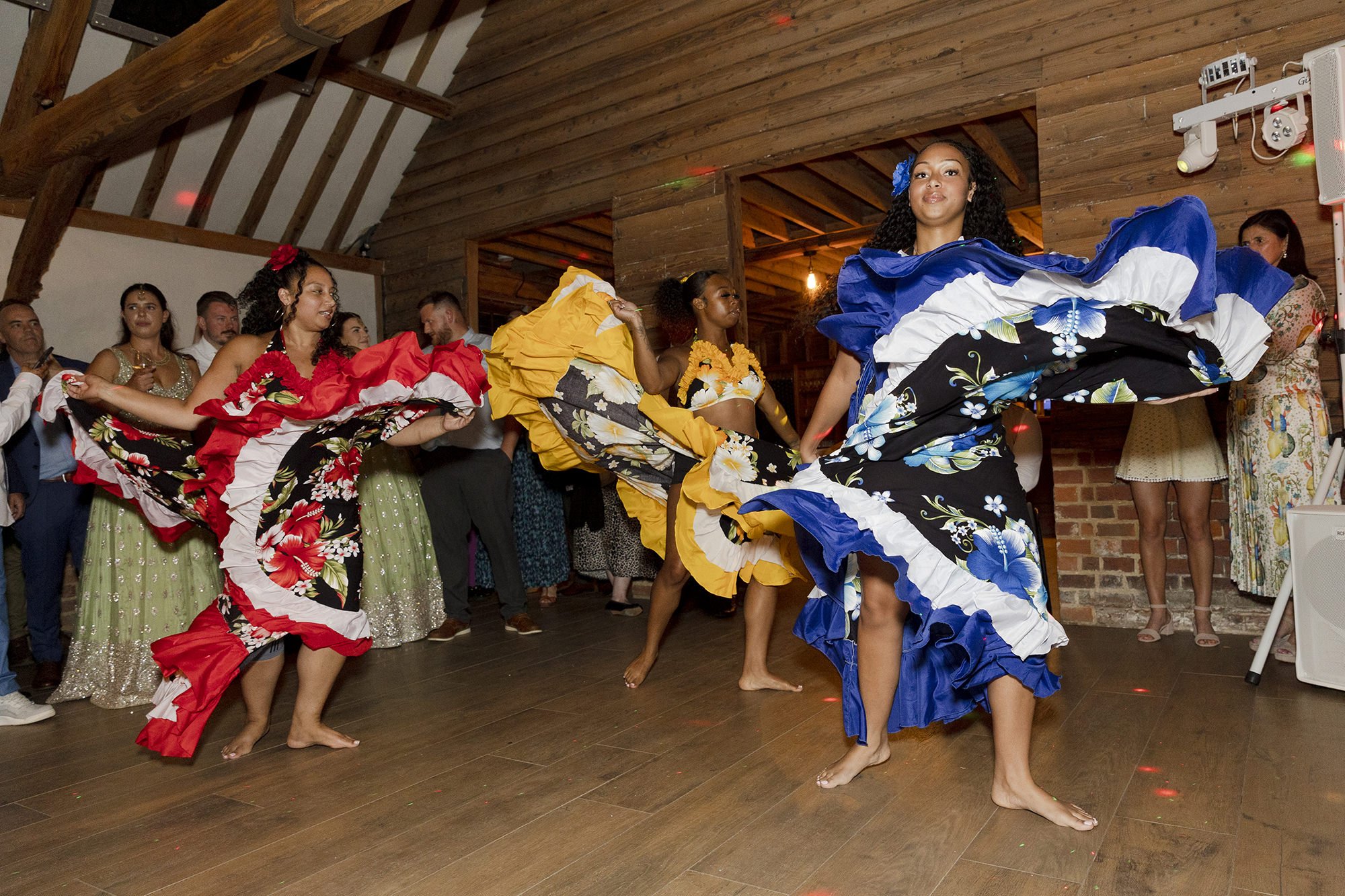 Women in colorful traditional dresses performing a dance at an indoor venue with wooden walls and people watching.