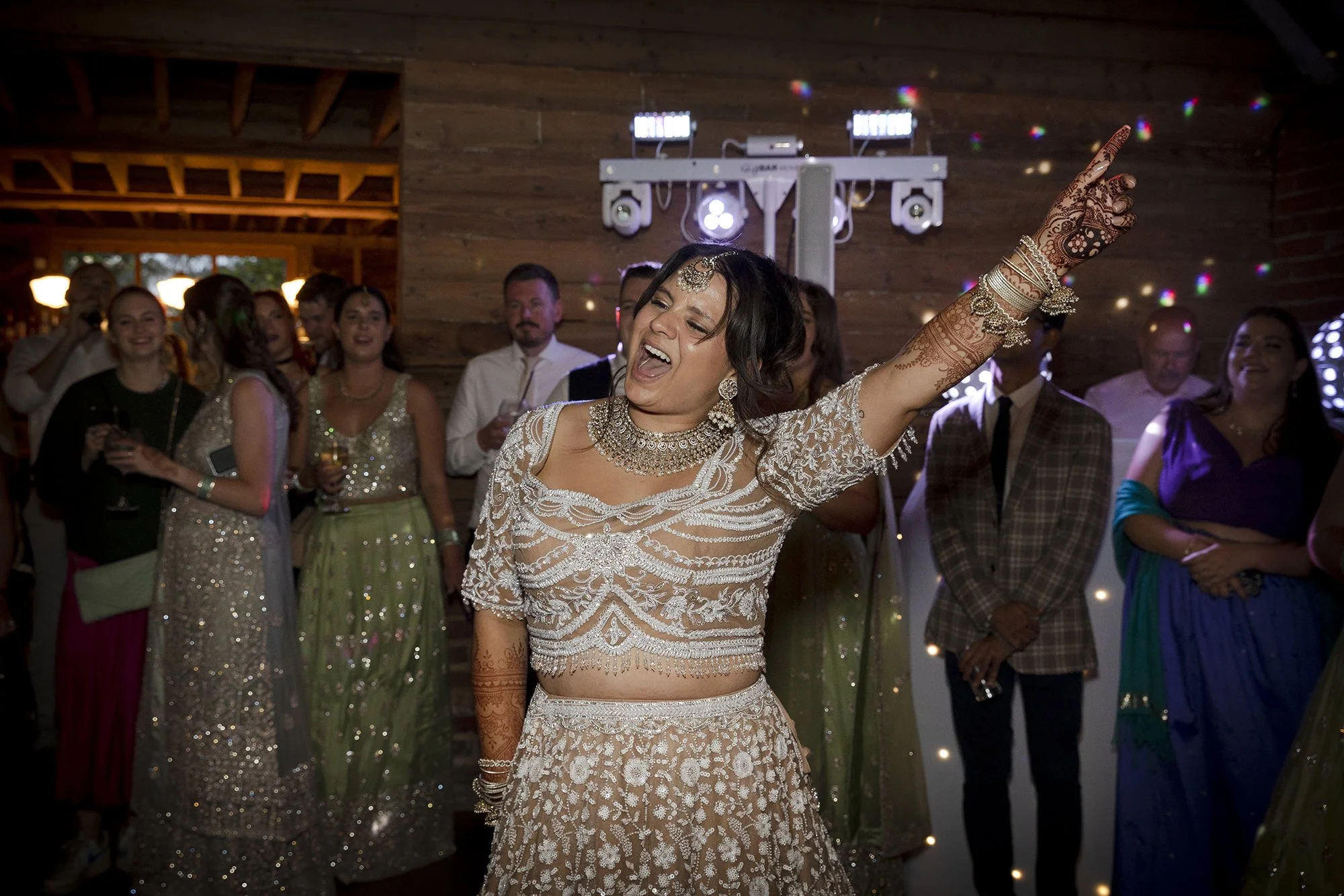 A woman in traditional Indian attire dancing at a celebration or wedding, surrounded by other people dressed in various colorful outfits, in a rustic indoor setting.