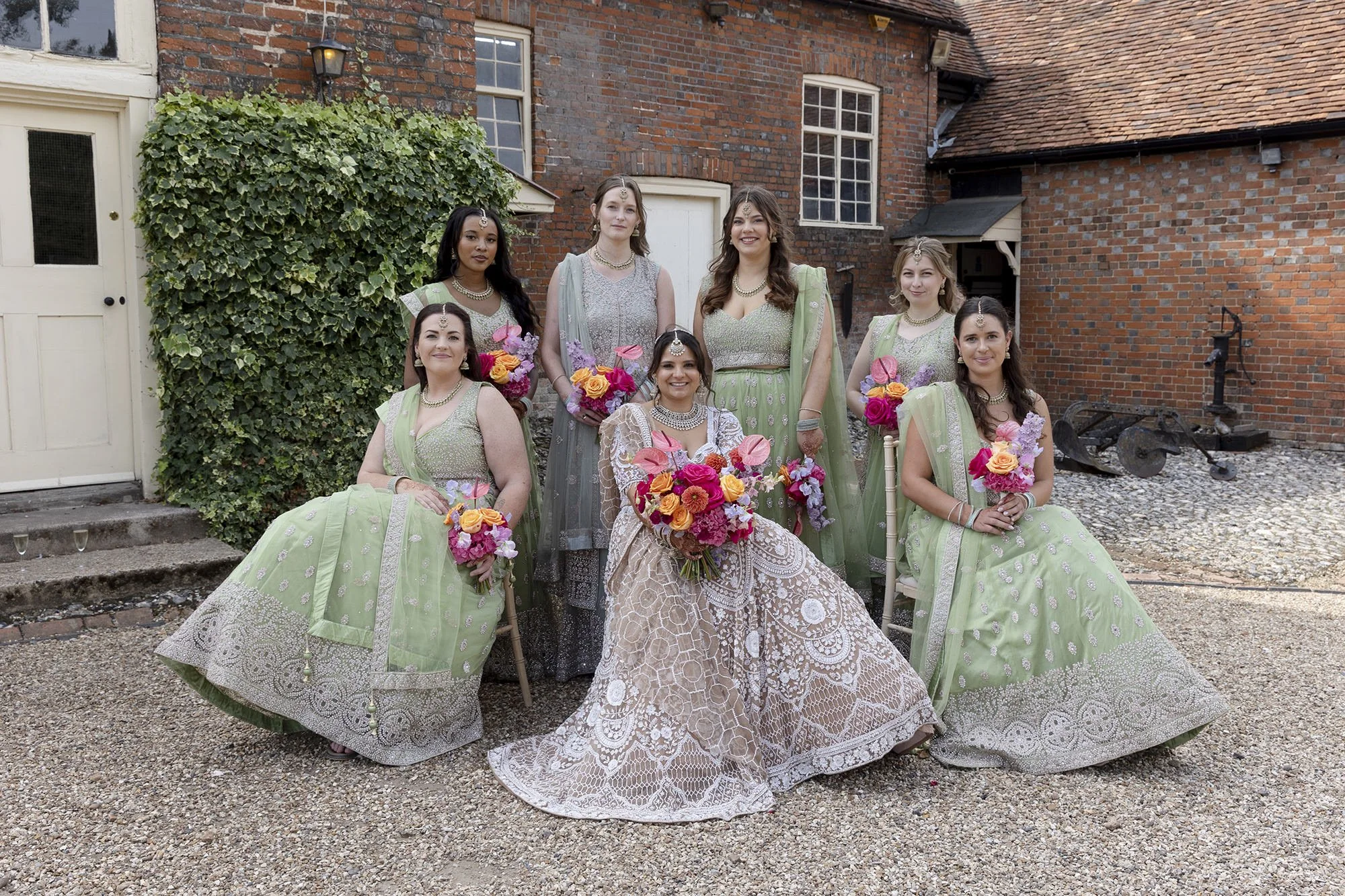 A group of nine women dressed in traditional Indian attire, holding floral bouquets, posing outdoors in front of a brick building with greenery.