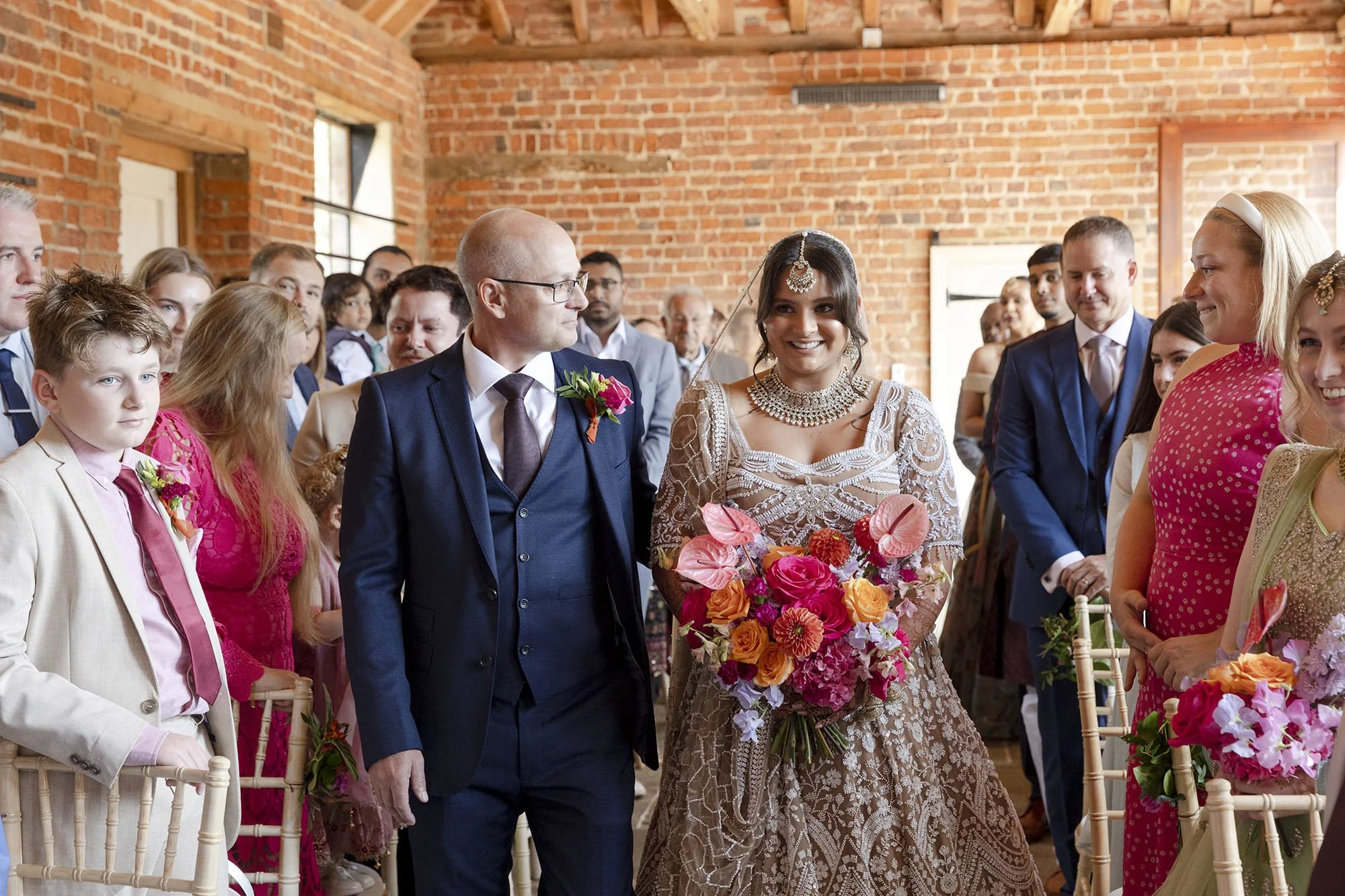 A bride and groom walking down the aisle during a wedding ceremony, surrounded by guests in a brick-walled venue.
