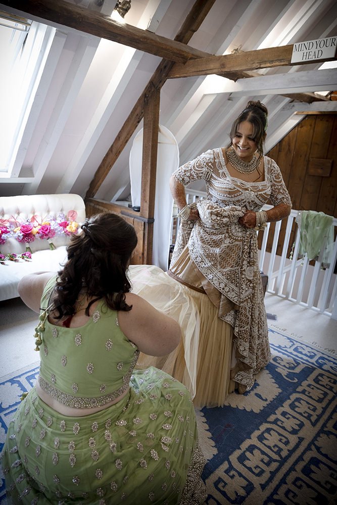Two women dressed in traditional Indian attire, one standing and smiling, the other sitting, in a cozy attic room with wooden beams and a floral decorated sofa.