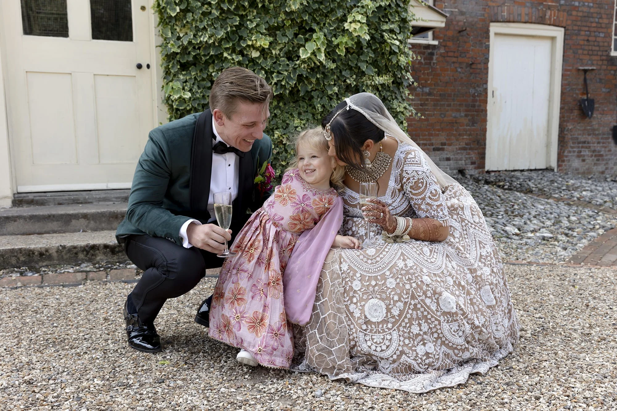 Bride and groom with a young girl, all smiling and holding champagne glasses, outdoors on a gravel surface near a brick building, with greenery behind.