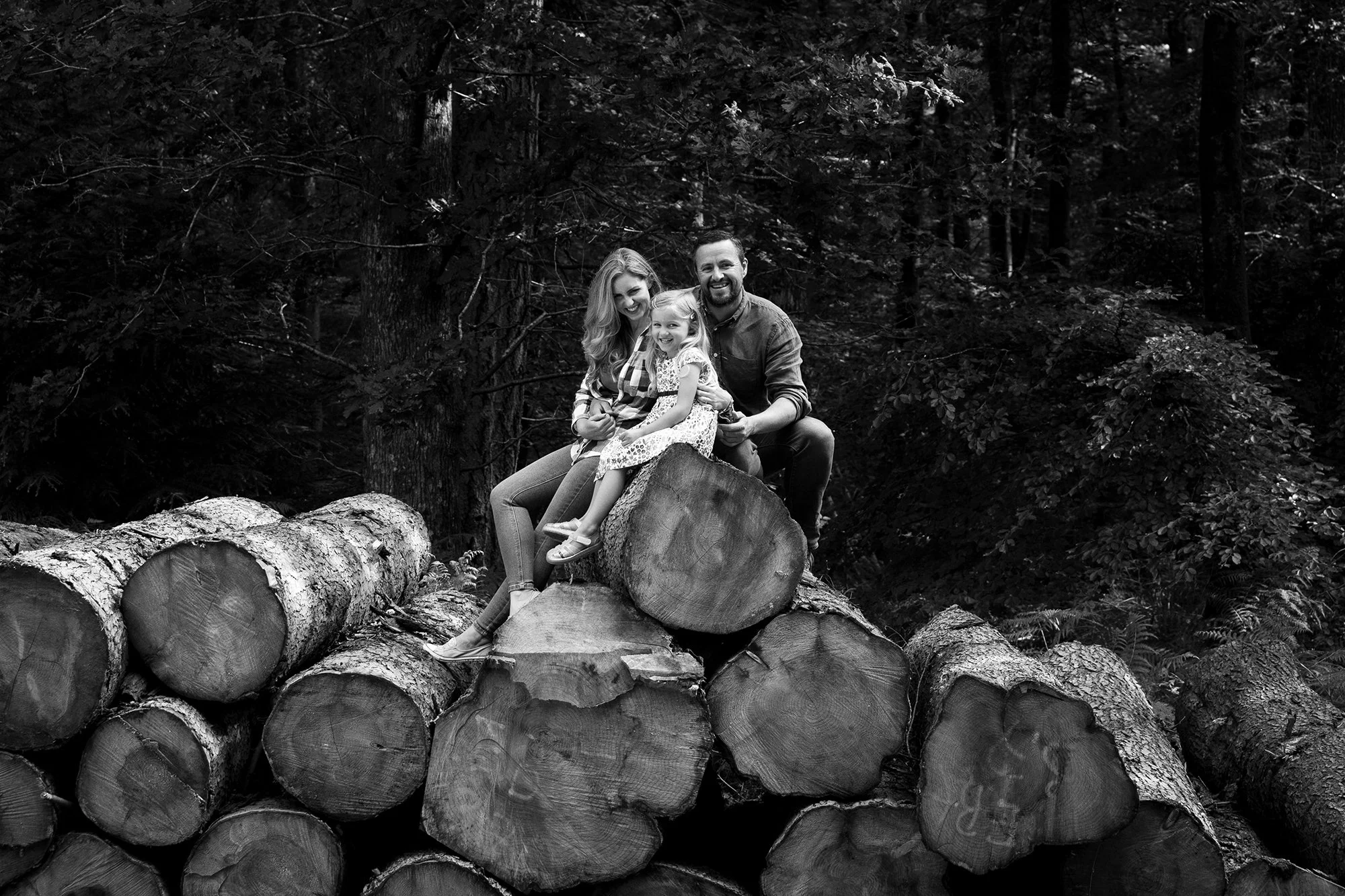 A family of three sitting on a pile of logs in a wooded area.