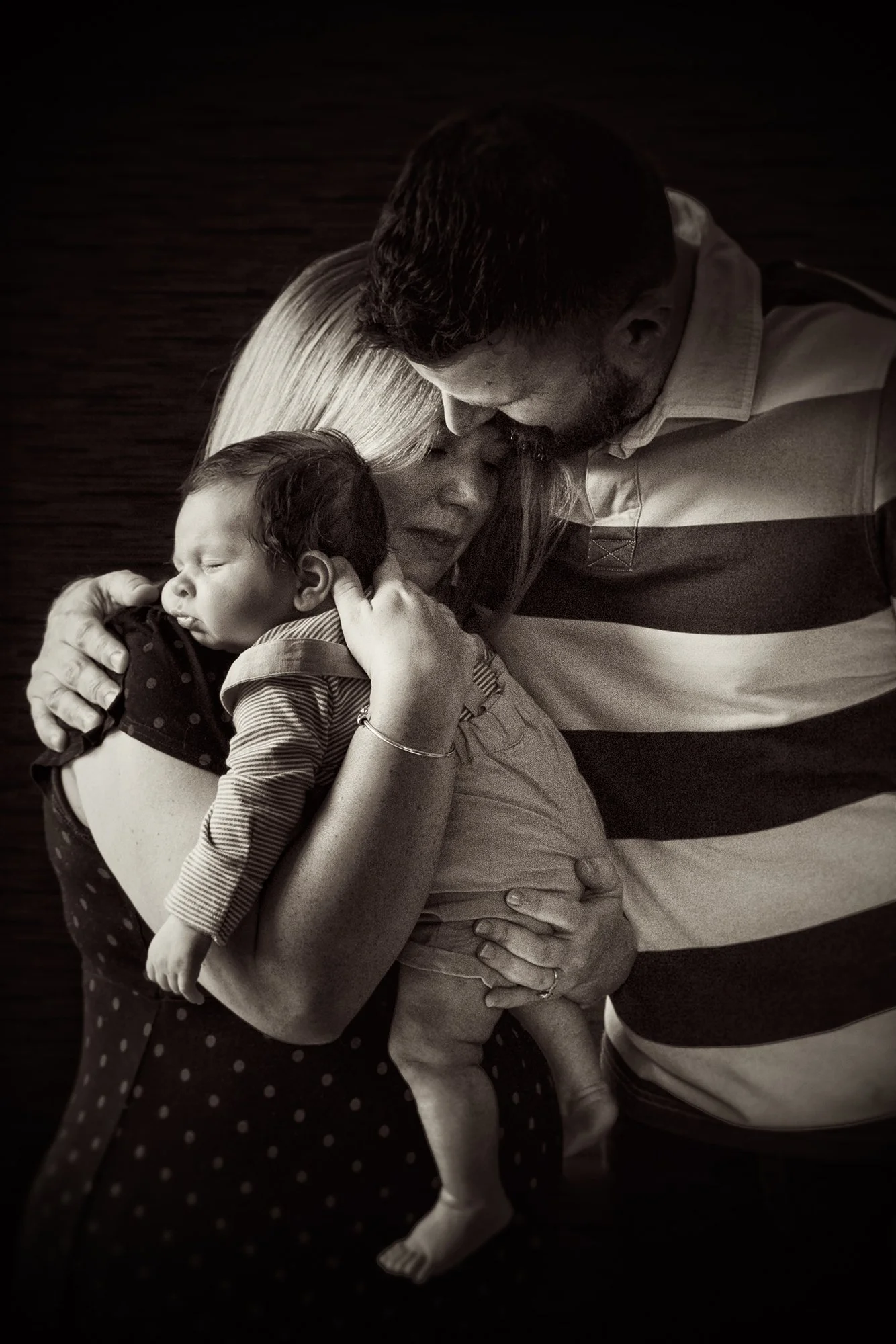 A family of three, including a woman, man, and a sleeping child, sharing a tender embrace in a close, intimate moment.