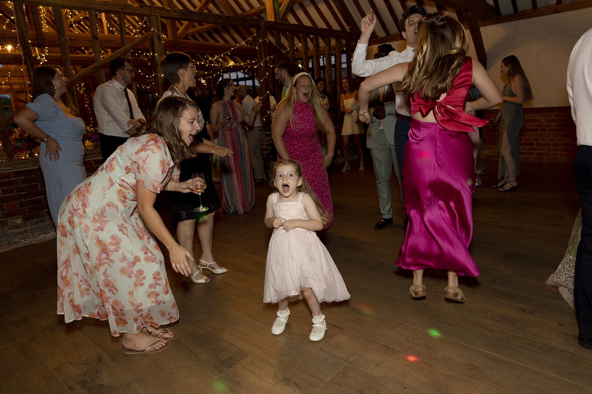 People dancing and celebrating at a party or wedding reception in a decorated venue with string lights. A young girl in a pink dress appears surprised or excited as she dances.