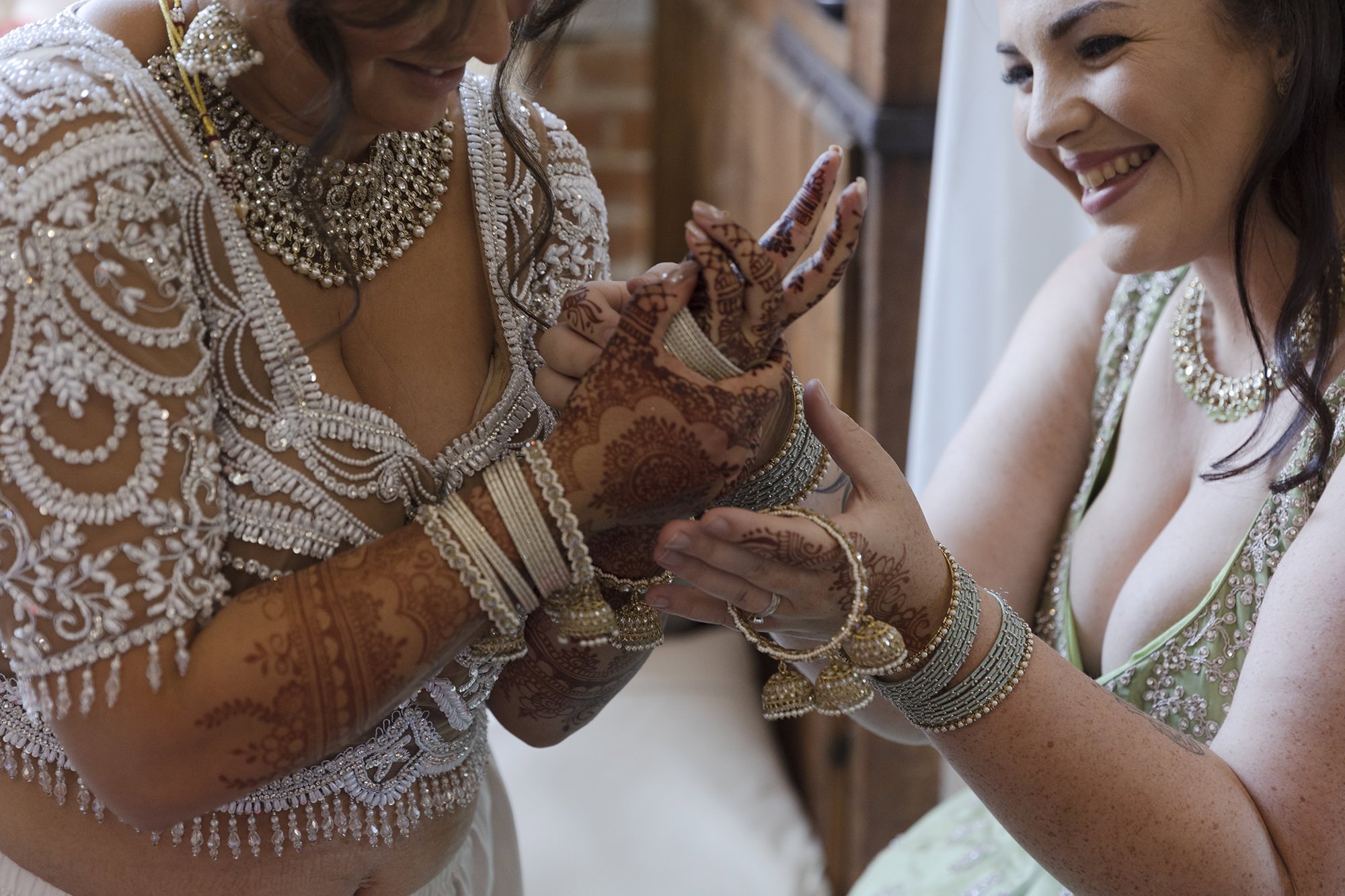 Two women celebrate a wedding, one wearing traditional Indian attire with intricate jewelry and henna on her hands, while the other wears a light green dress and silver jewelry. They are smiling and holding hands.