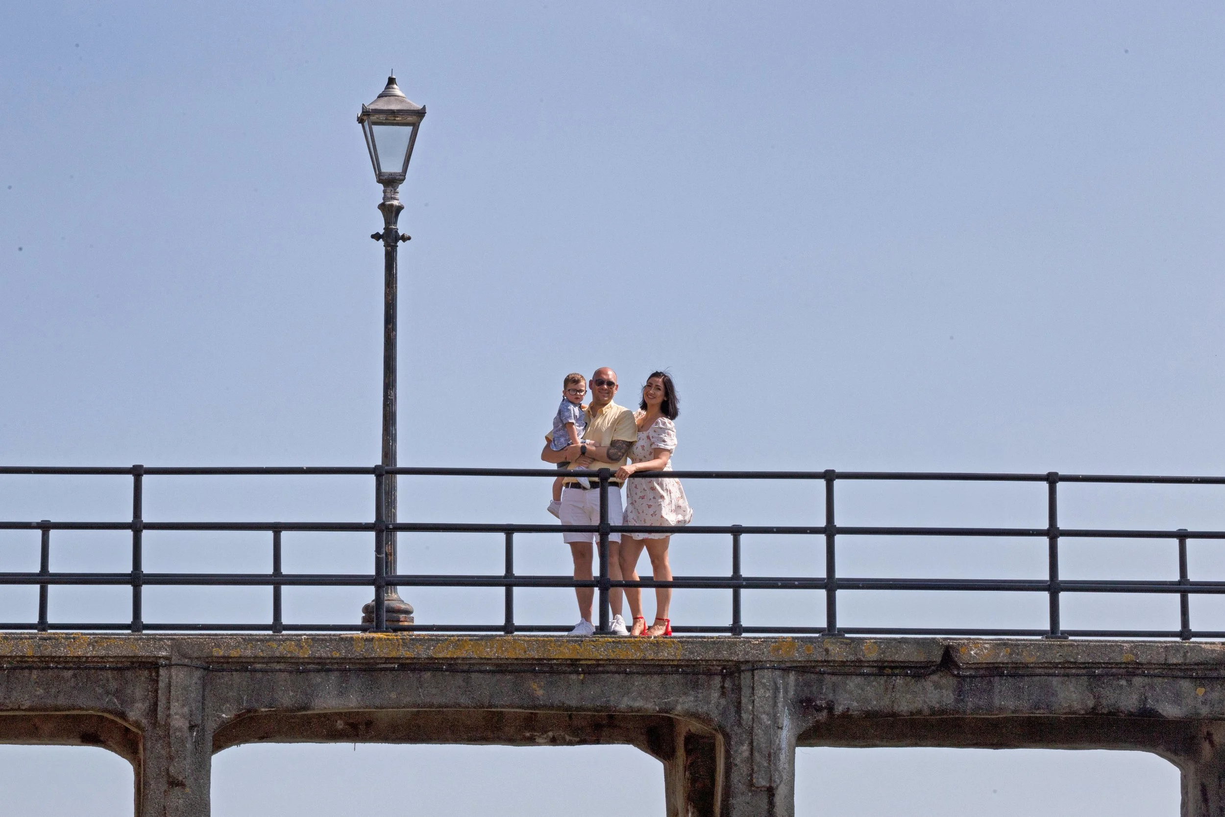 A family of three standing on a bridge with a rail, near a lamppost, against a mostly clear blue sky, smiling and looking at the camera.