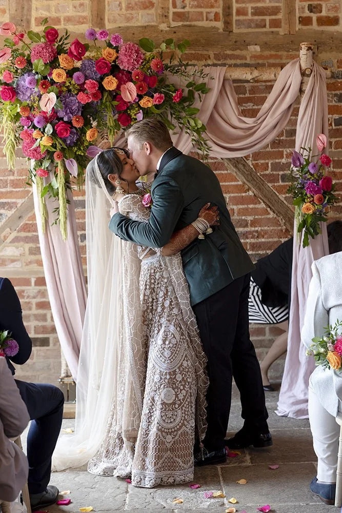 A bride and groom kiss during their wedding ceremony under a floral arch with pink, purple, and orange flowers beside a brick wall.