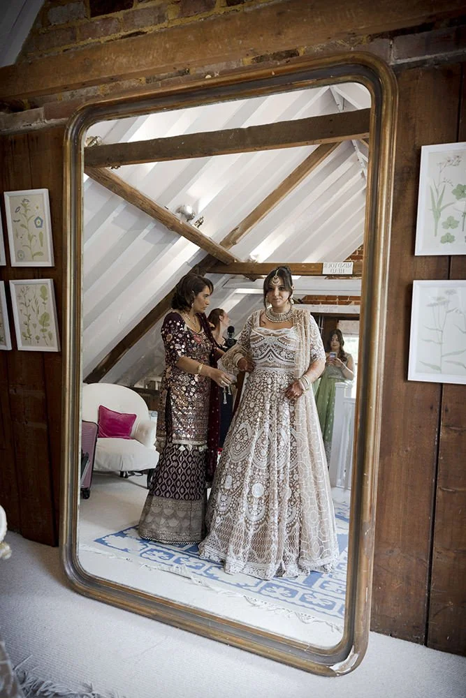 Reflected in a large ornate mirror, a bride in an elaborate cream-colored wedding gown stands with a woman holding a microphone, both dressed in traditional South Asian attire. Two women in the background, one in green, are taking photos. The setting