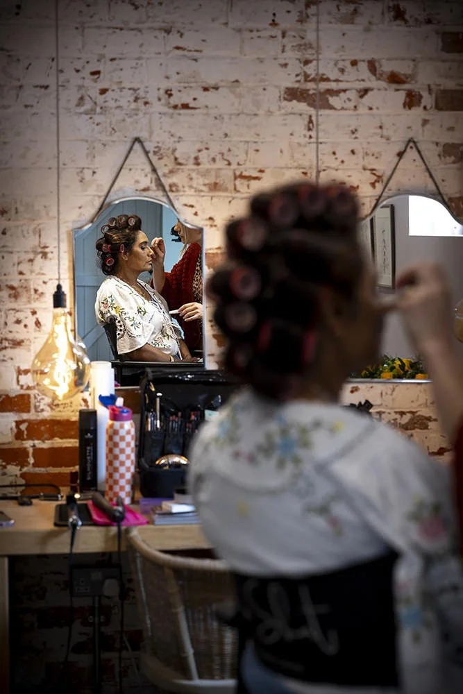 A woman getting her makeup done in a salon, with the reflection of another woman in a mirror, sitting in a chair with hair rollers