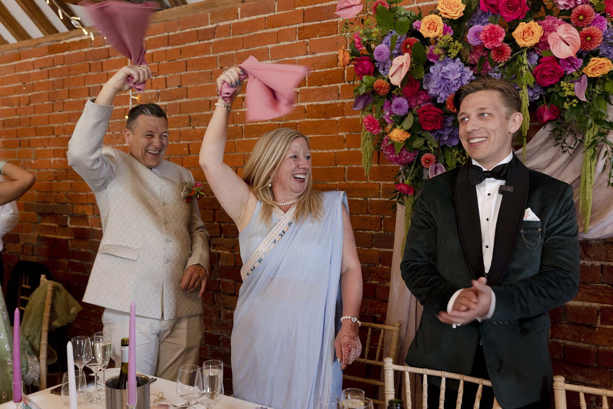 Group of people celebrating at a wedding reception. A woman in a blue saree is holding pink napkins, smiling, and dancing. A man in black tuxedo is standing nearby, smiling. A man in traditional cream-colored attire is also smiling and holding pink n