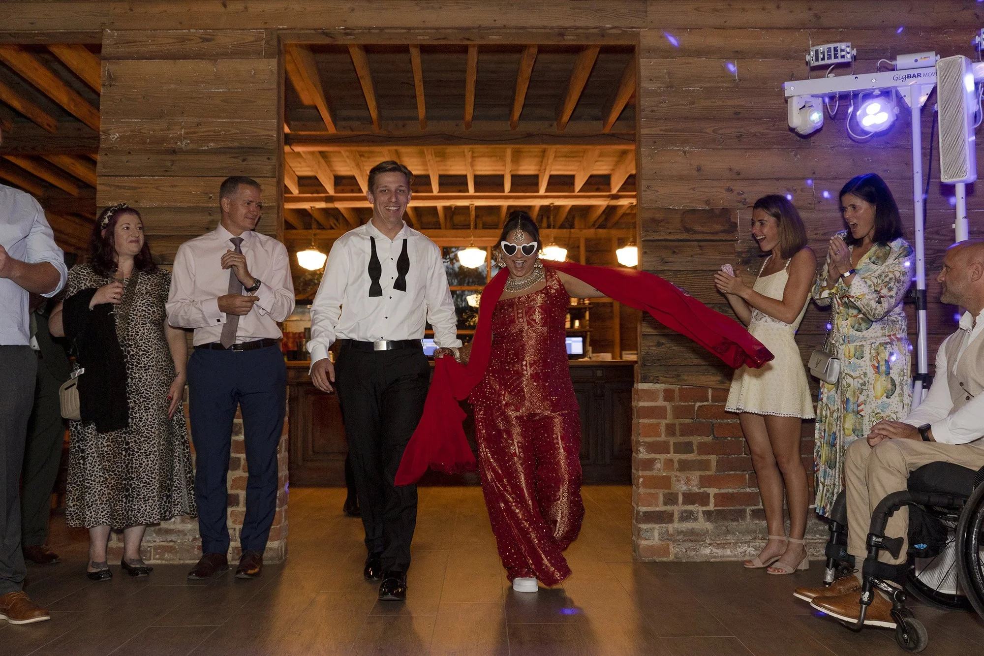 People at a celebration, with a woman in a red dress and sunglasses, walking confidently with a red cape, surrounded by friends or guests in a cozy, rustic venue with wooden walls and ceiling.