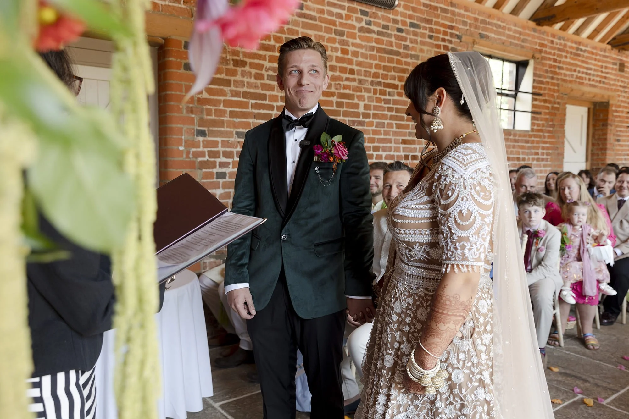 A wedding ceremony with a groom, bride, and officiant inside a brick building. The groom wears a tuxedo with a boutonnière, and the bride wears a lace dress with henna on her hands, jewelry, and a veil. The guests sit in the background, smiling at th