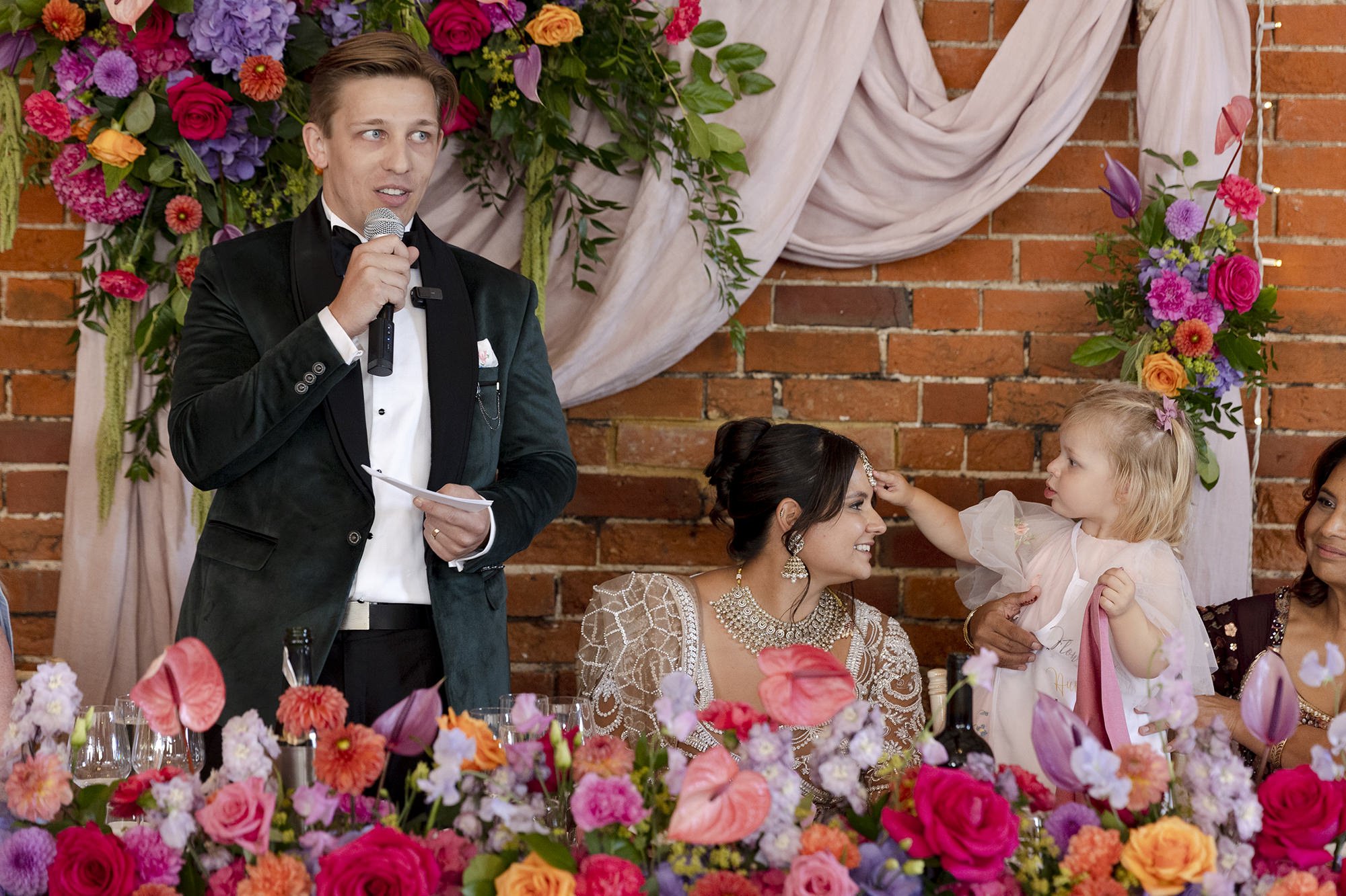A man in a tuxedo giving a speech at a wedding reception, standing next to a bride and a young girl, surrounded by colorful flowers and brick walls.