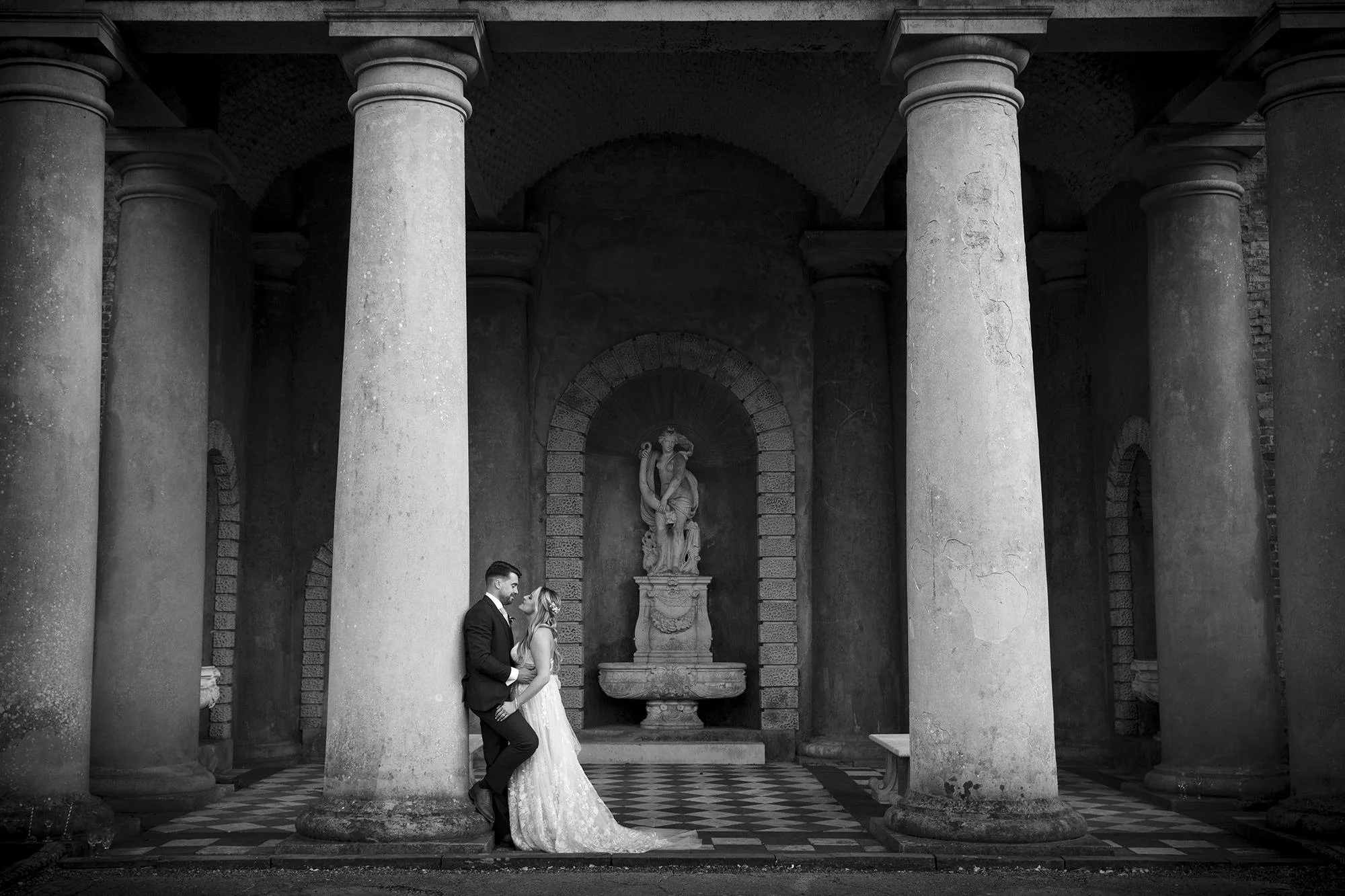 Couple posing in wedding attire in front of large columns and a statue, black and white photo.