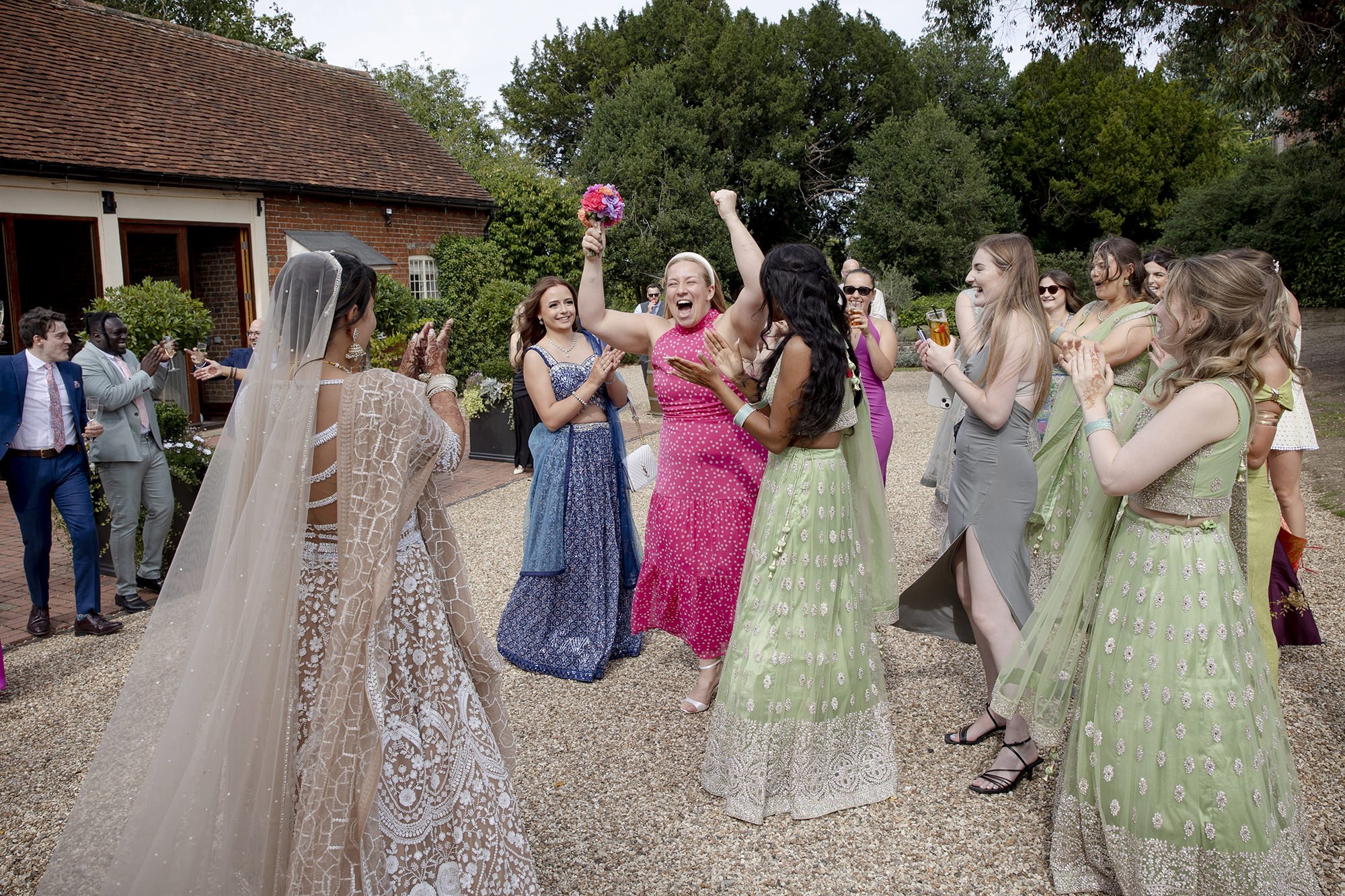 A group of women and men celebrating outdoors at a wedding, with the bride in a gown and veil, and other women dressed in colorful Indian-style dresses. One woman in a pink dress is happy and throwing her hands up. The guests are smiling, clapping, a