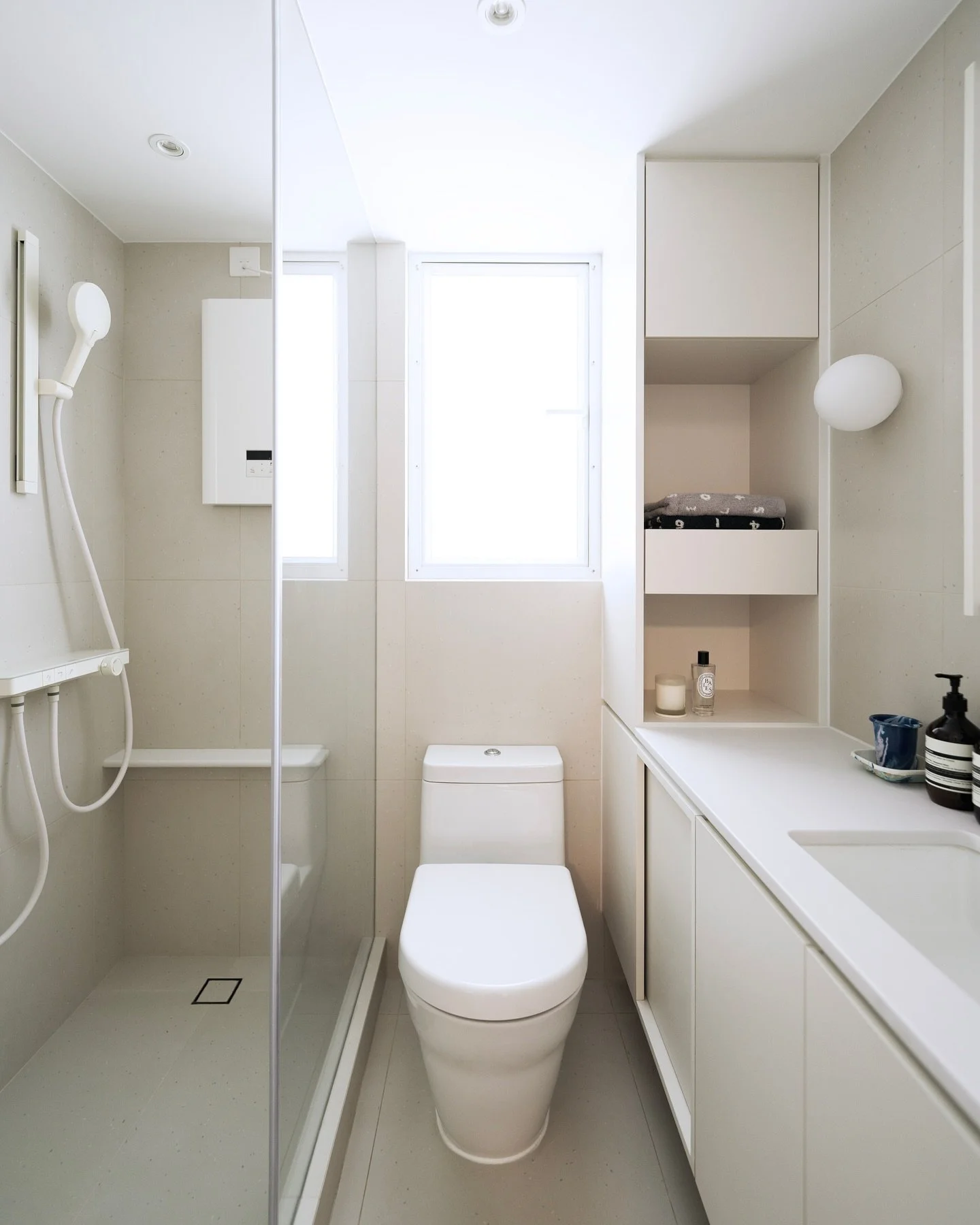 -
Residential project: 
實用面積: 430sqft

我們將客廳的流動性設計思維巧妙融入衛浴空間，以暖灰色系霧面瓷磚鋪陳基底，搭配白色立體幾何造型衛浴配件，透過材質冷暖對比與線條變化，在極簡框架中構築豐富的觸覺層次體驗。

針對有限空間的收納需求，在馬桶側打造頂天立地式窄身儲物高櫃，採用開放層架結合隱藏儲格的複合設計，既滿足毛巾、衛浴備品的多元收納，通透的設計更有效消弭小空間的壓迫感。同步在地櫃門片導入滑軌式趟門設計，精準控制開合幅度，將每寸收納效能極大化。

+
如有興