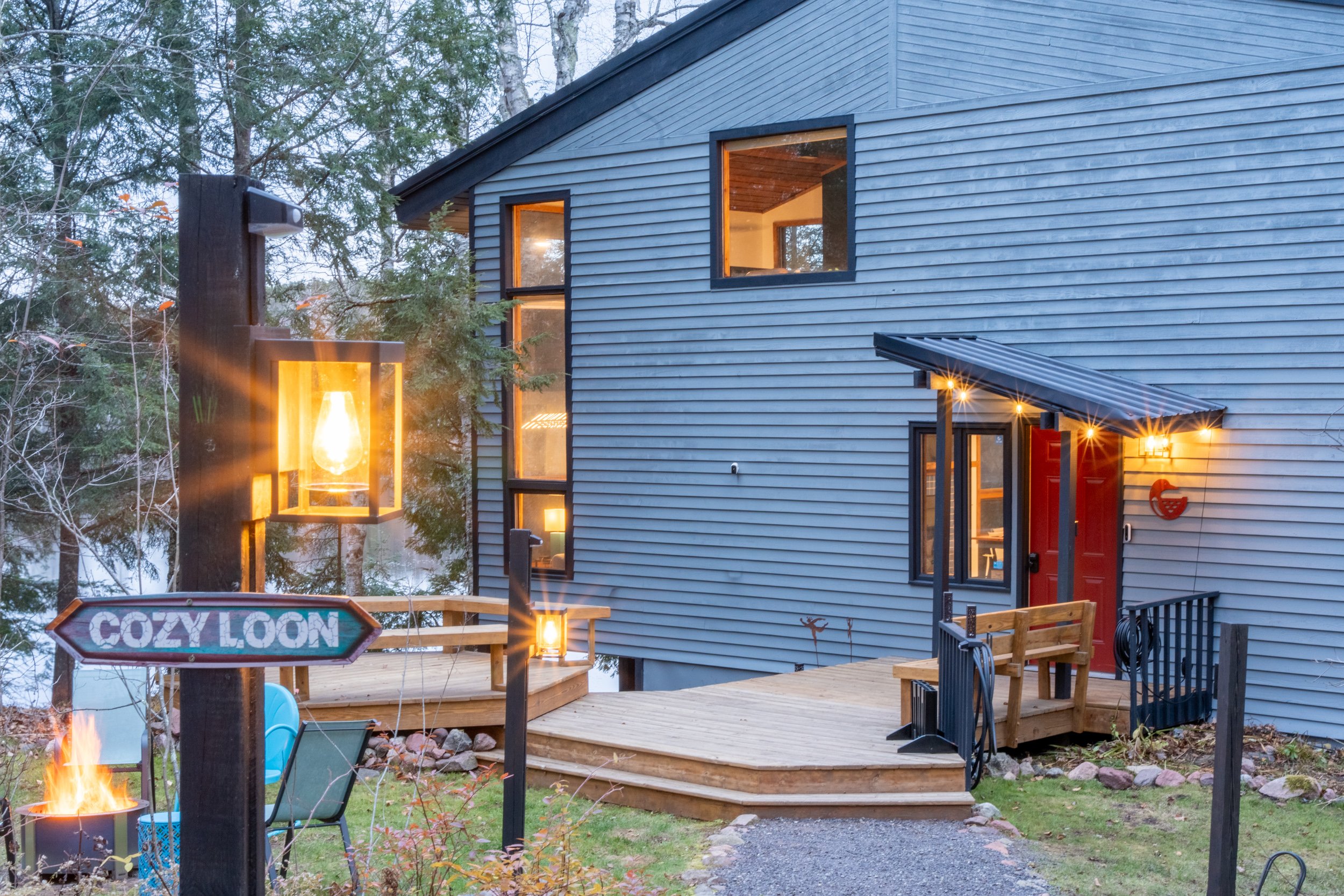 A modern two-story house with blue siding, illuminated windows, and a small red door, surrounded by trees and outdoor lighting. A wooden deck extends from the house with a fire pit and outdoor furniture. There is a sign that says "COZY LOON" in the y