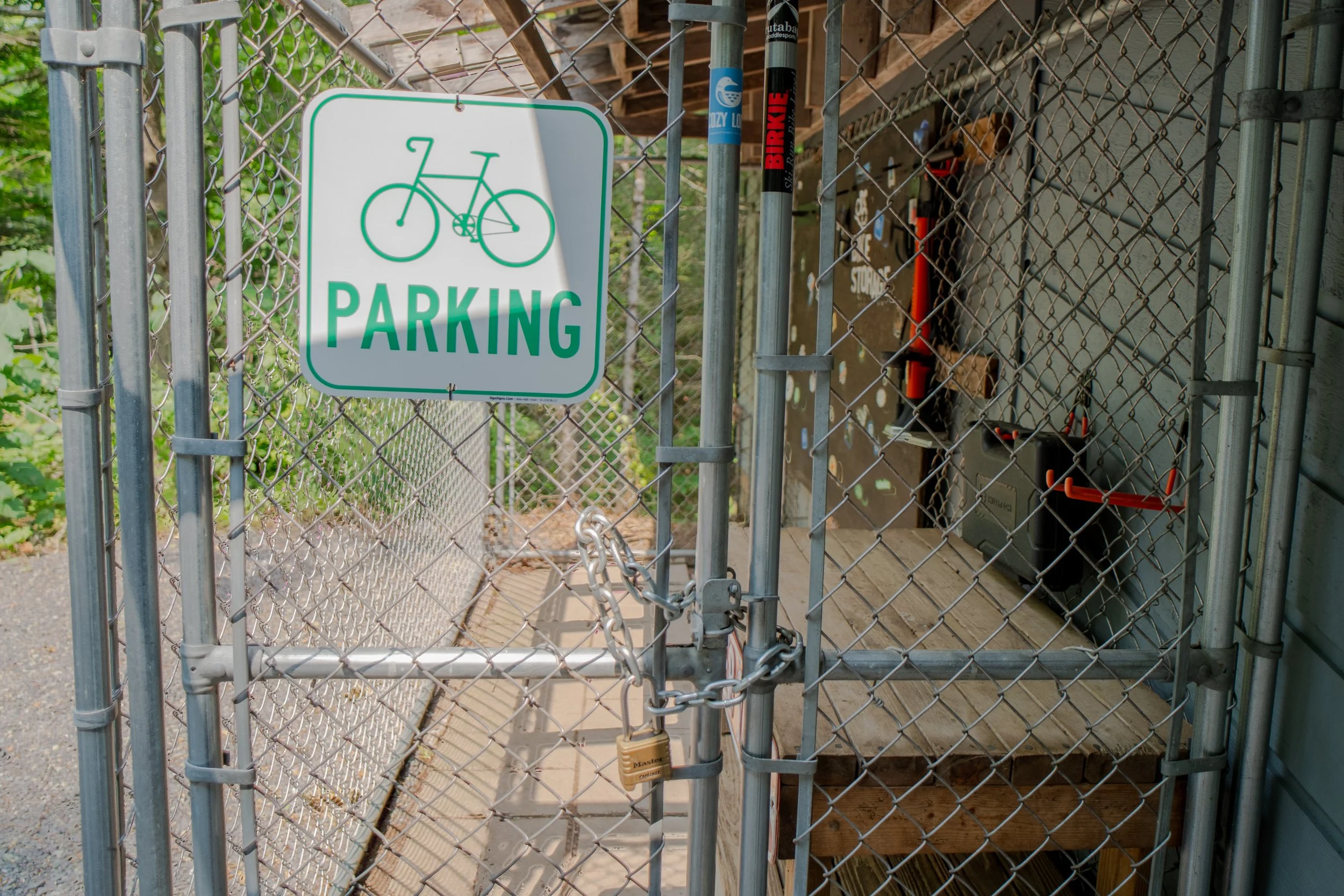 A metal gate with a white sign that has a green border and green text reading 'PARKING' along with a bicycle icon in green. The gate is secured with a chain and lock, leading to a wooden deck area with tools and equipment visible behind the fence.