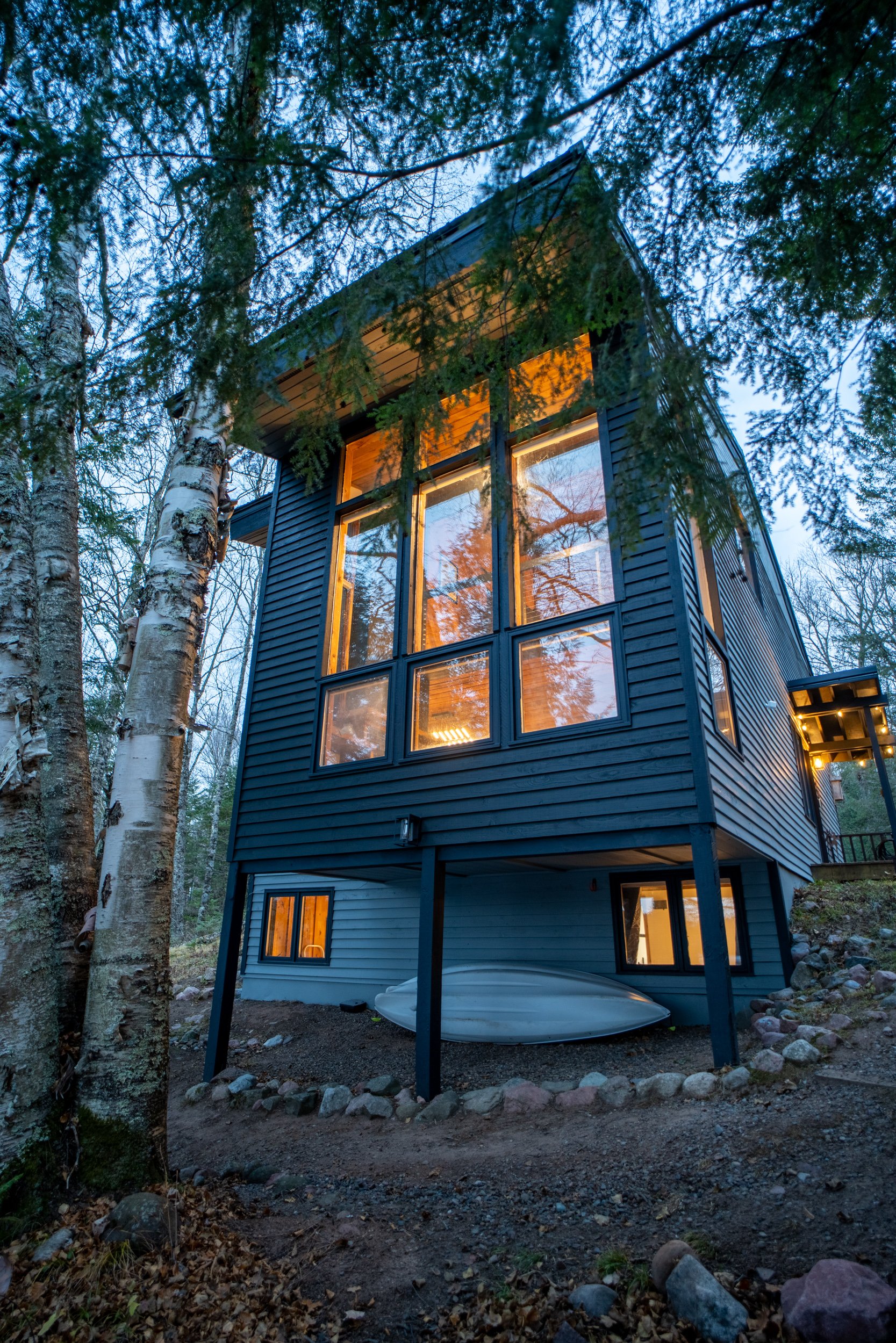 A modern black house with large windows situated in a wooded area with trees around it, lit from inside during dusk.
