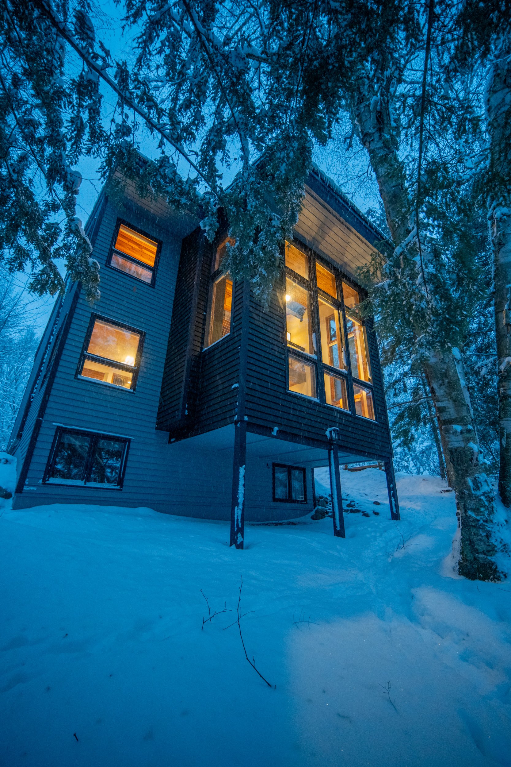 A modern multi-story house illuminated from within, situated among snow-covered trees during a winter evening.