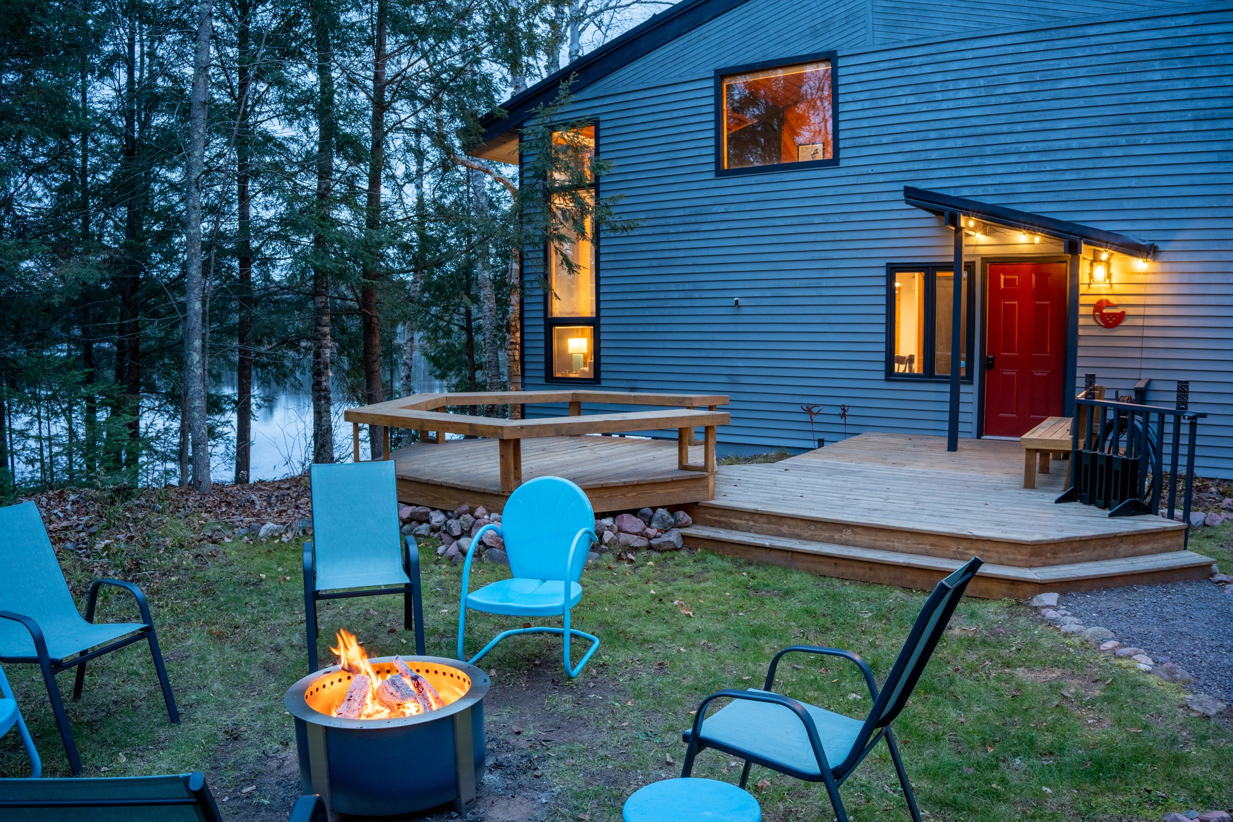 A house with blue siding and large windows is shown in the woods at dusk. The house has an outdoor deck with chairs and a fire pit burning in the yard. The scene is illuminated by interior and exterior lights.