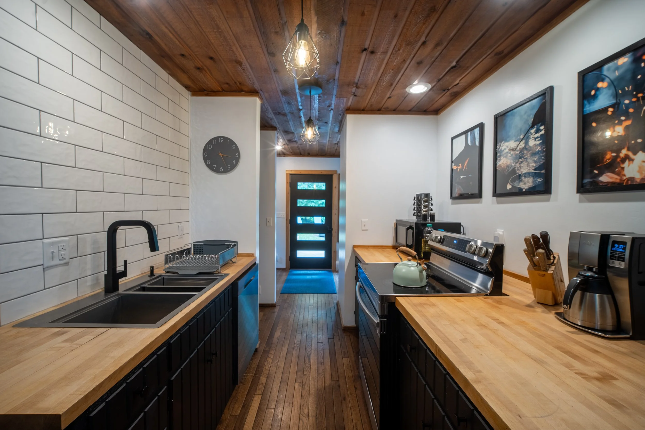 Modern kitchen with white subway tile walls, wooden ceiling, black cabinets, and a kitchen island. Includes a clock on the wall, a stainless steel stove, and framed pictures on the right wall.