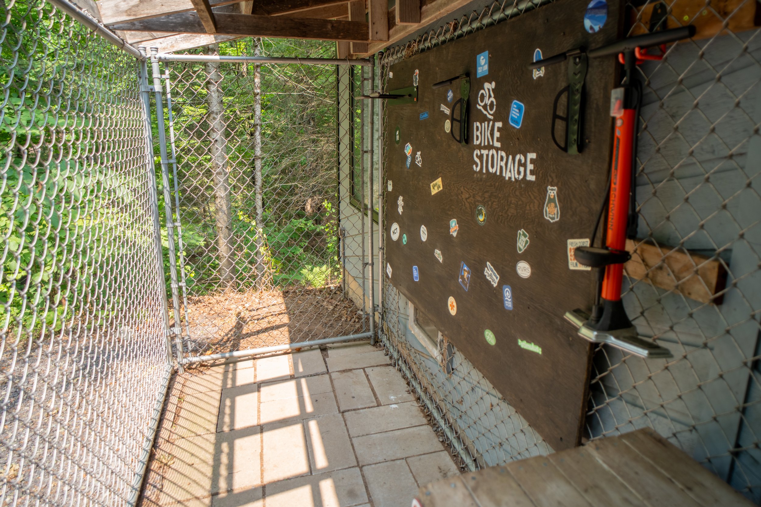 A fenced outdoor area with a wooden bike storage board decorated with stickers, attached to a chain-link fence, with trees and foliage in the background.