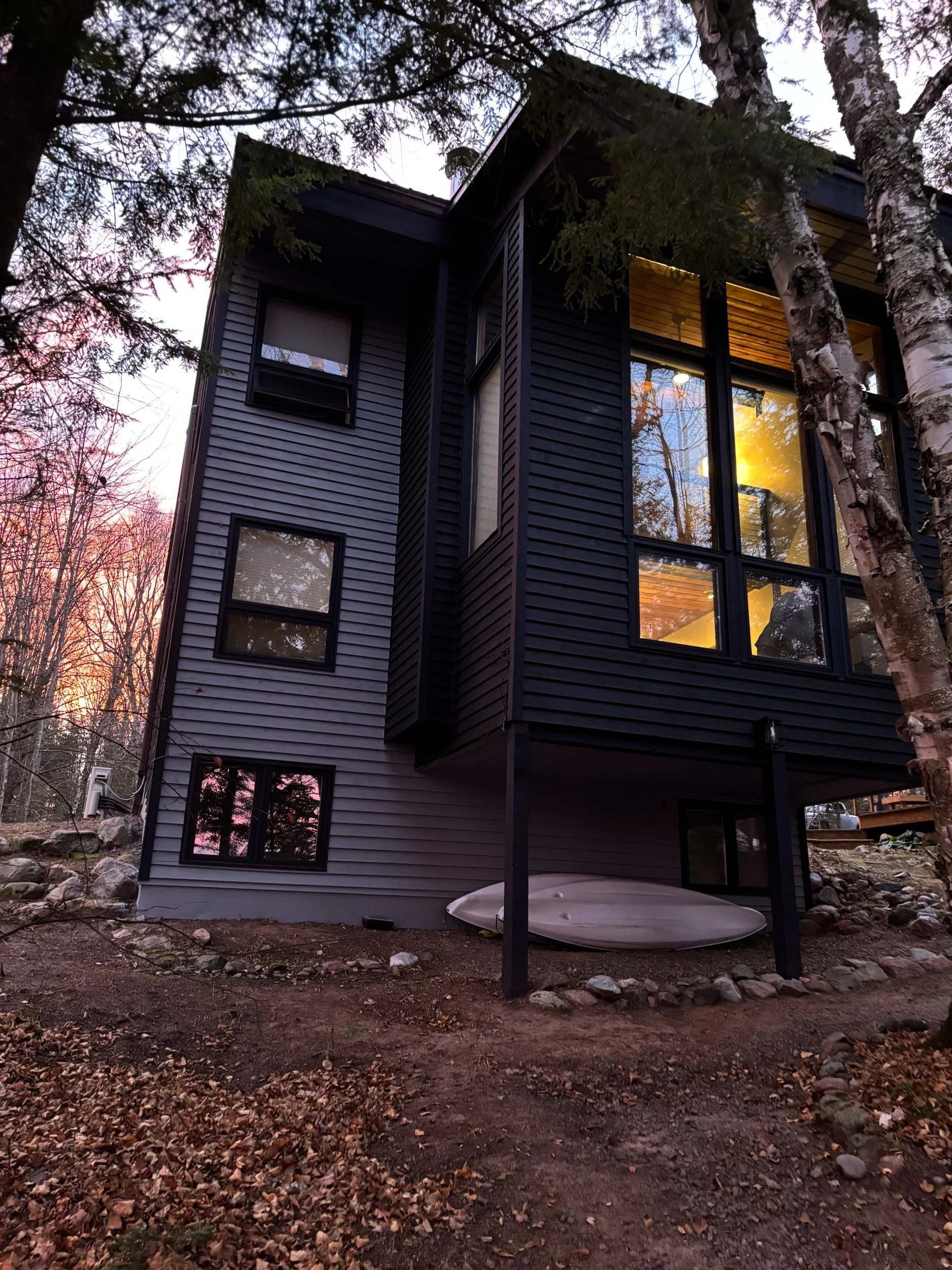 Image of a modern black house surrounded by trees during sunset, with large windows reflecting the sky and interior lights.