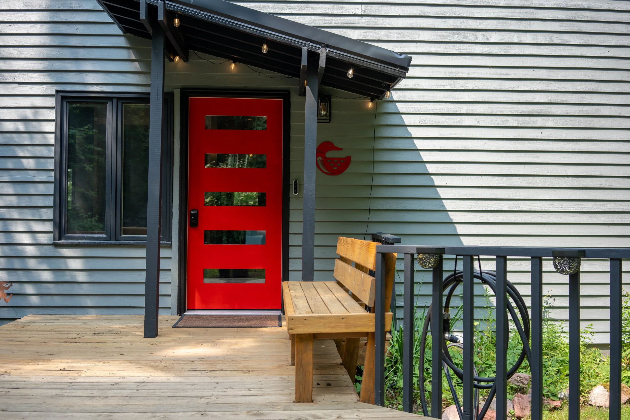 A house exterior with a red front door with horizontal window panels, a shuttered window, and a small porch with bench seating and string lights hanging from the porch roof.