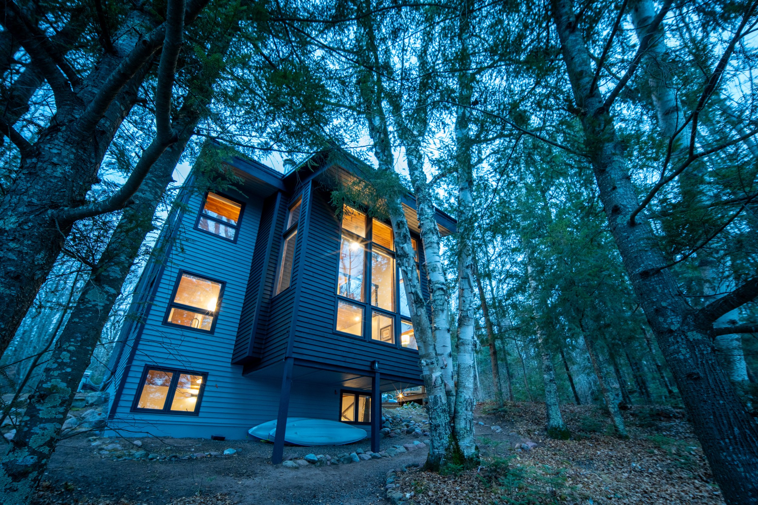 A modern blue house with large windows is surrounded by tall trees in a wooded area, illuminated from within.