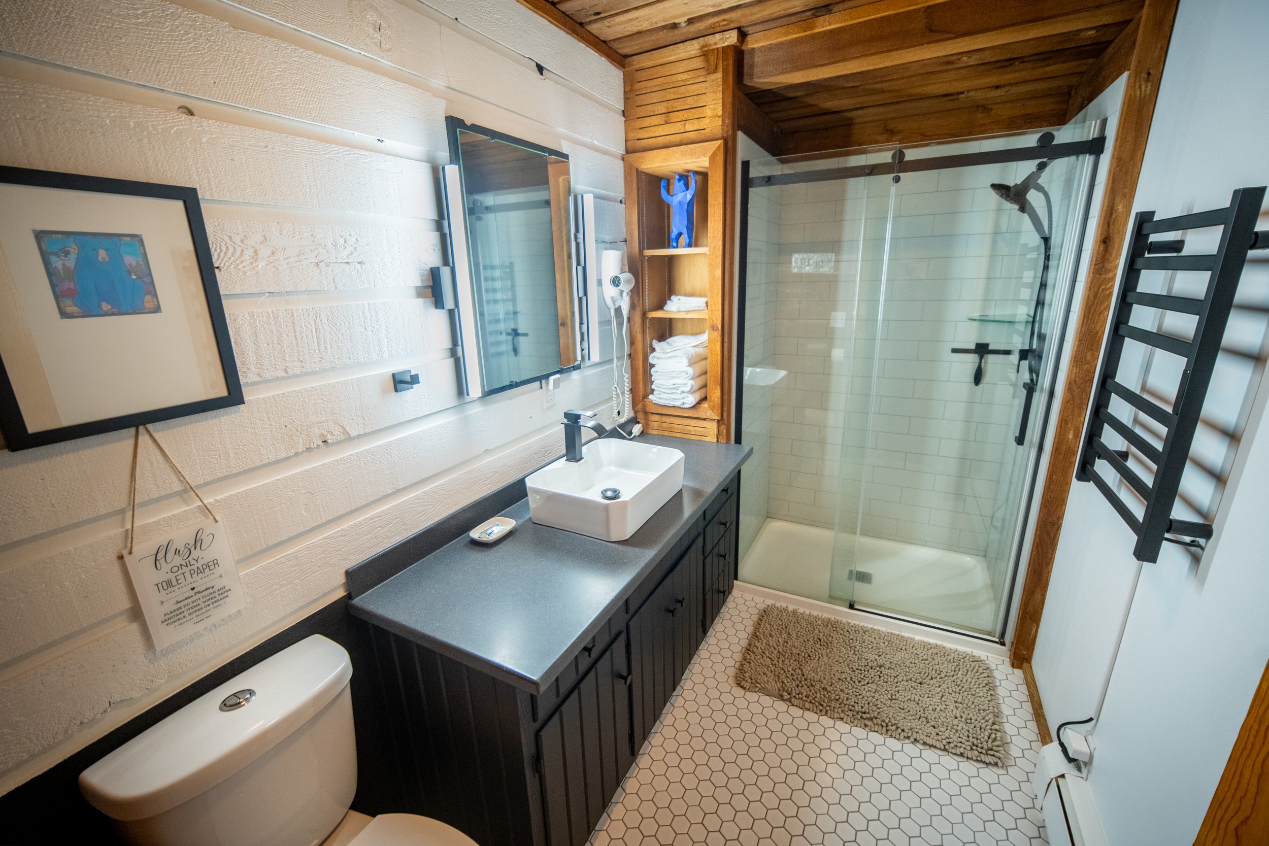 A bathroom with a white toilet, black vanity with a white sink, and a glass-enclosed shower with the door open. The wall is decorated with framed artwork and a sign that reads 'flush only toilet paper'. There is a wooden shelf with towels and a blue 