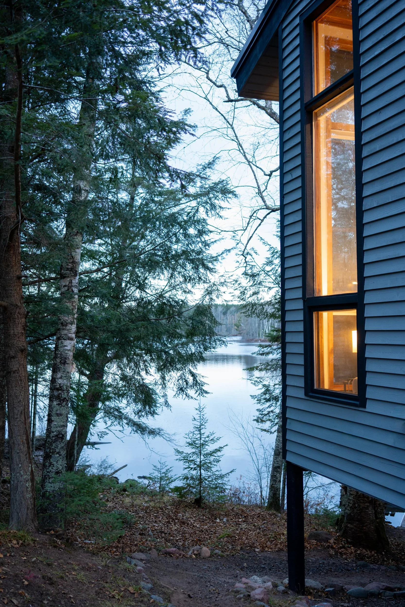 A house with dark exterior siding and large windows with warm interior lighting, located next to a wooded area with tall trees and over a frozen or partially frozen lake during dusk or early evening.