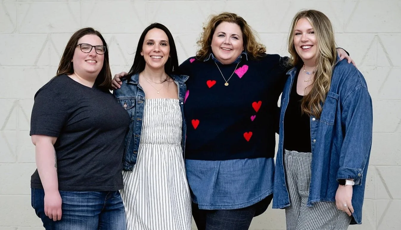 The Reclaim Therapy team. Four trauma therapists near me standing close together, smiling, with one woman's arm around the others, against a beige brick wall.
