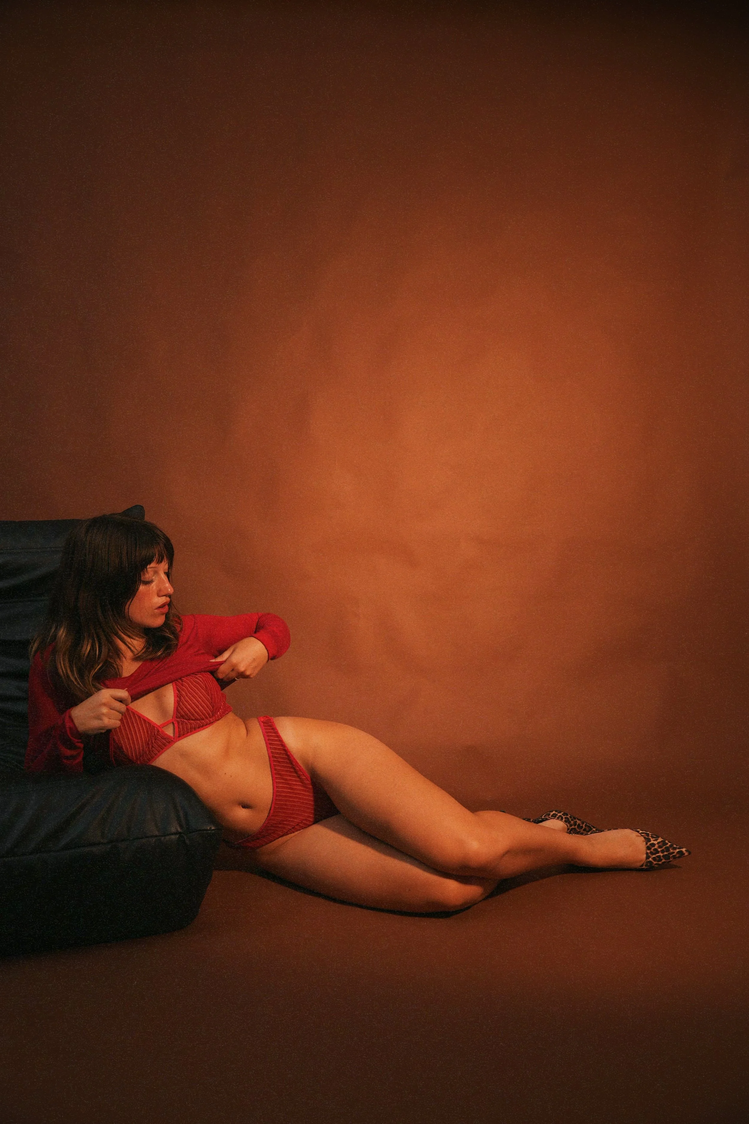Une femme assise sur un canapé noir, portant un ensemble de lingerie rouge et des chaussures à motif léopard, posant contre un mur marron.