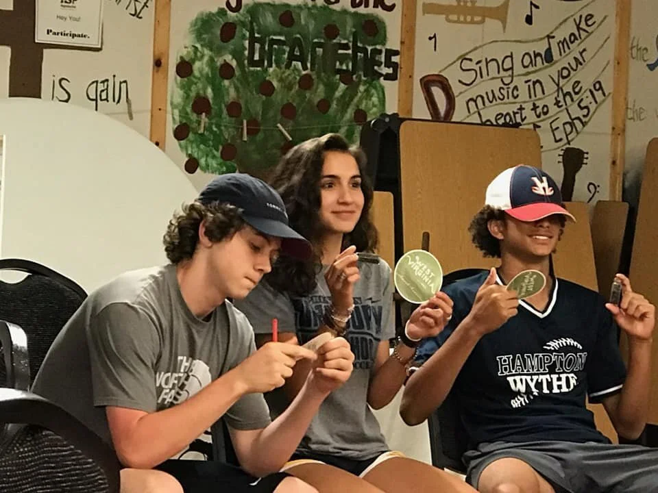 Three teenagers sitting in a room, smiling. One boy is wearing a gray T-shirt and a cap, engrossed in a small piece of paper. A girl in the middle has long, curly hair, and she is smiling at the camera. The girl on the right is wearing a dark sports jersey, a cap, and is holding a round green sticker and a small object, giving a thumbs-up.