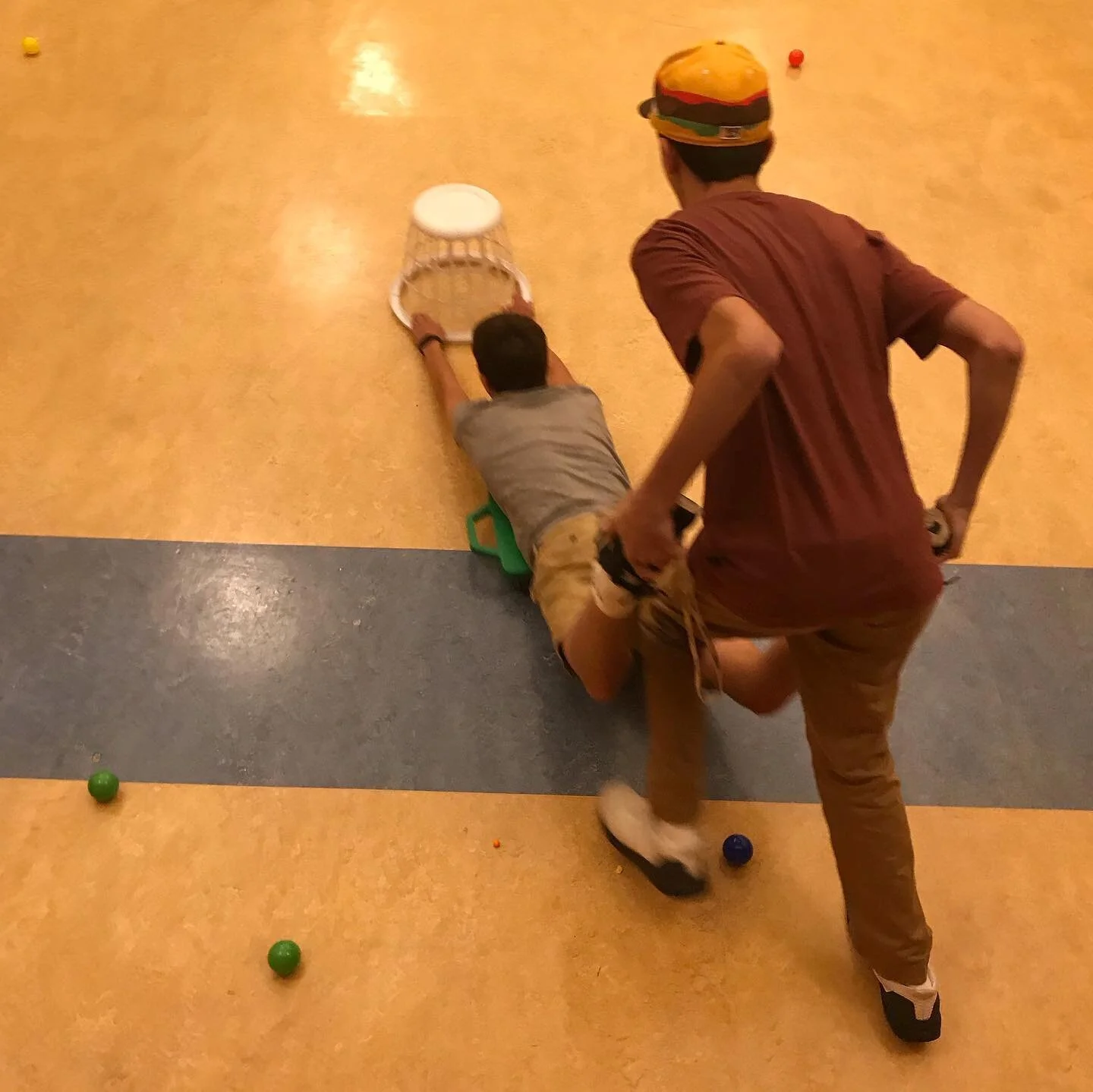 Two boys playing a game of croquet, with one lying on the ground aiming a mallet at the ball and the other standing nearby on a colorful indoor floor.