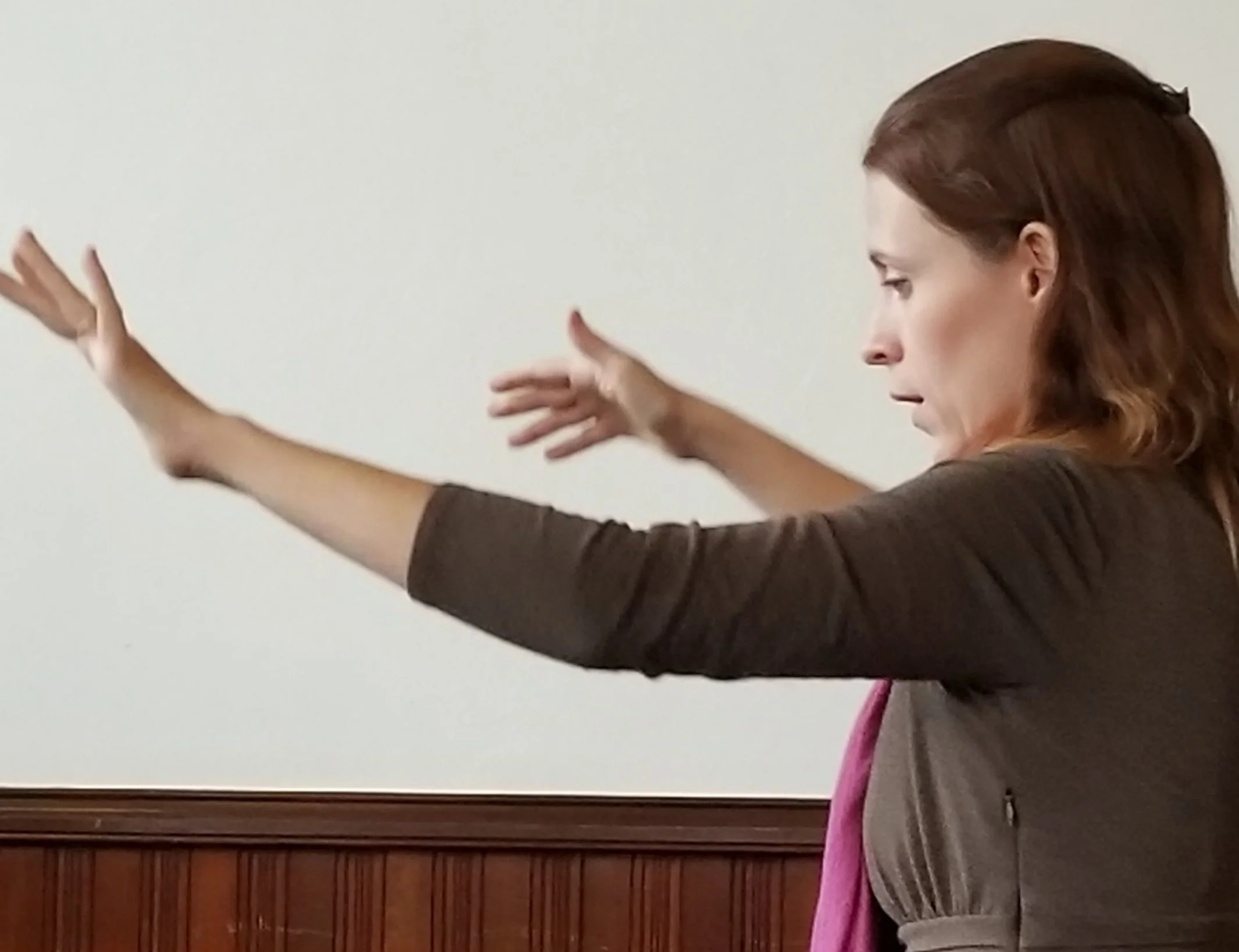 A woman with brown hair extending her arms outward in a defensive or expressive gesture, standing in front of a plain white wall with wood paneling at the bottom.