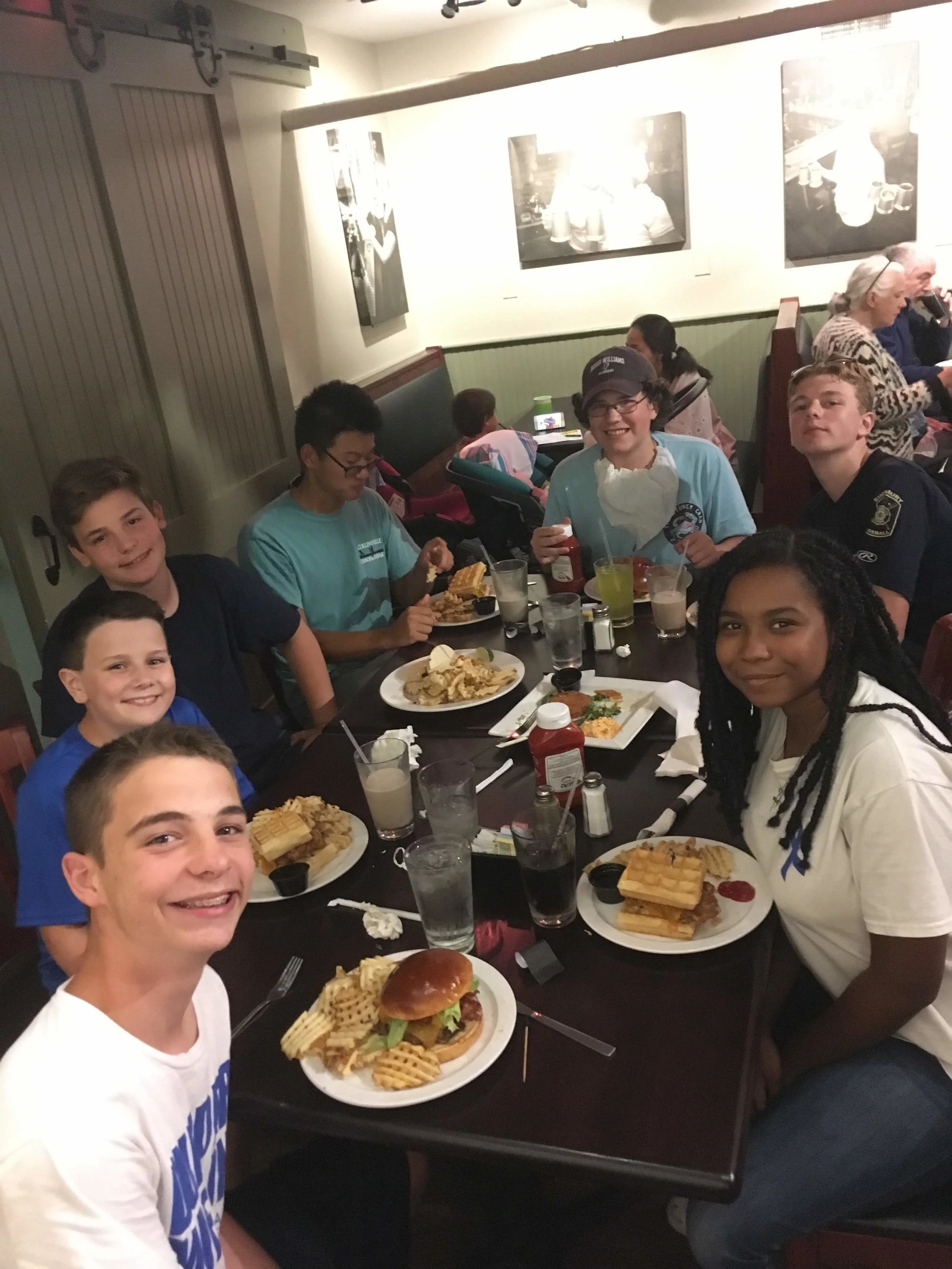 A group of children and teenagers sitting around a restaurant table with plates of food, including waffles, burger, and drinks, smiling and enjoying their meal.