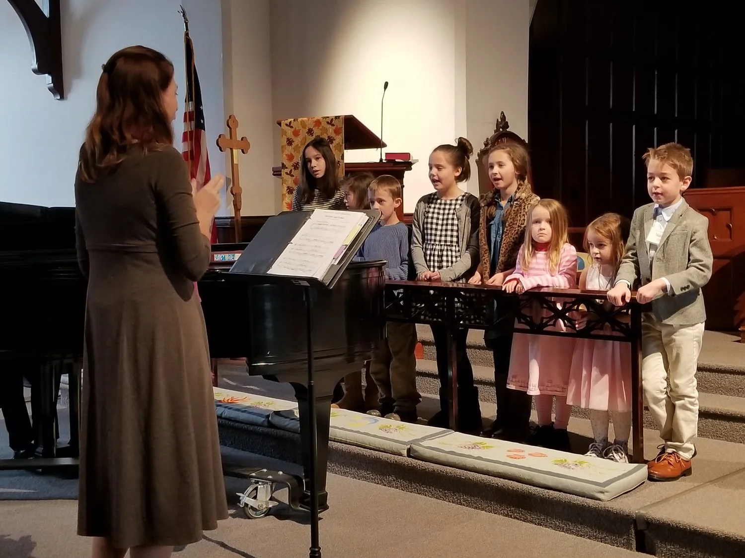 A woman directing a children's choir in a church. The children are standing behind a music stand and singing, with some on a small platform. The scene includes a piano and a church setting with an American flag in the background.