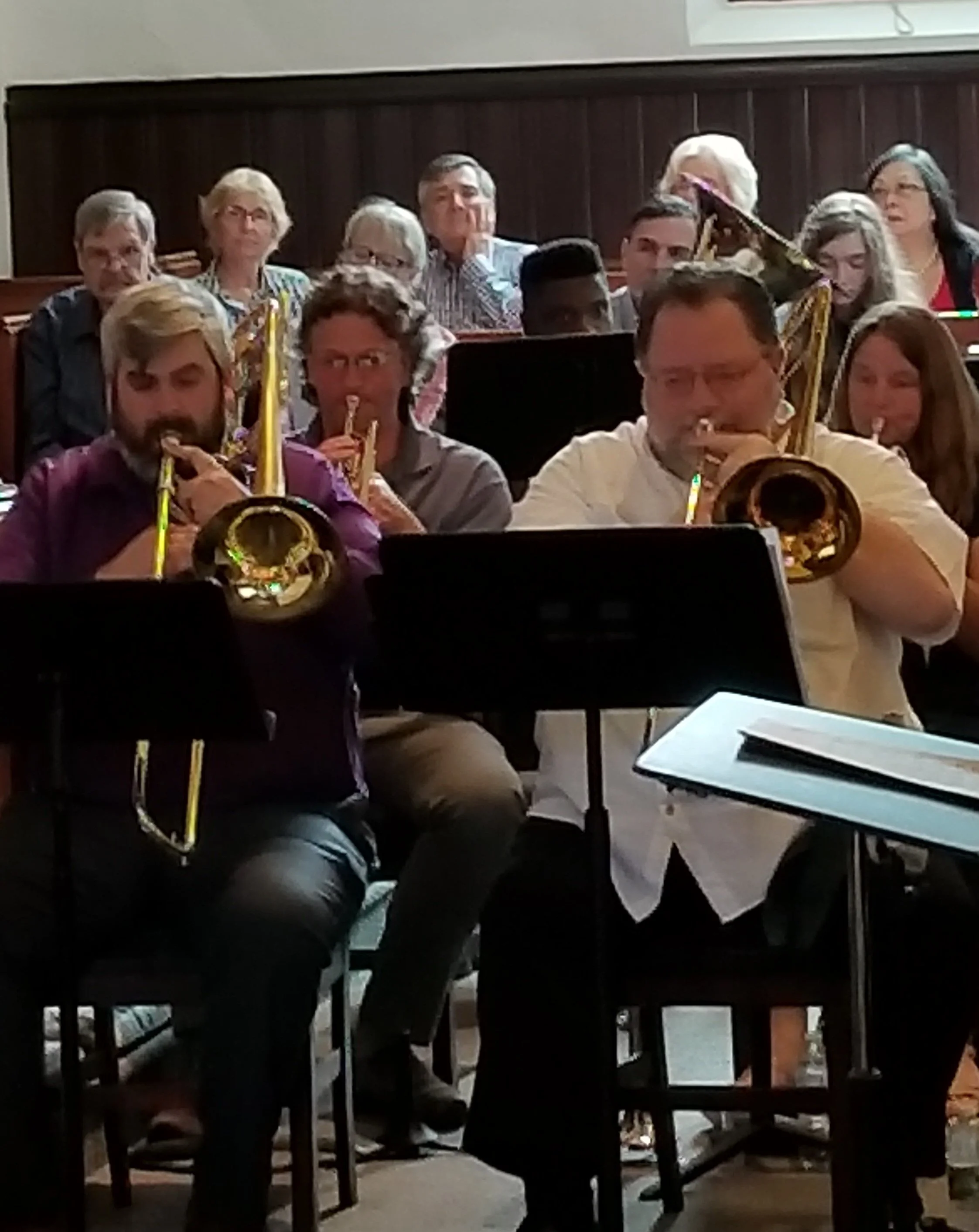 An ensemble of musicians playing brass instruments, seated in front of music stands, with some audience members seated behind them.