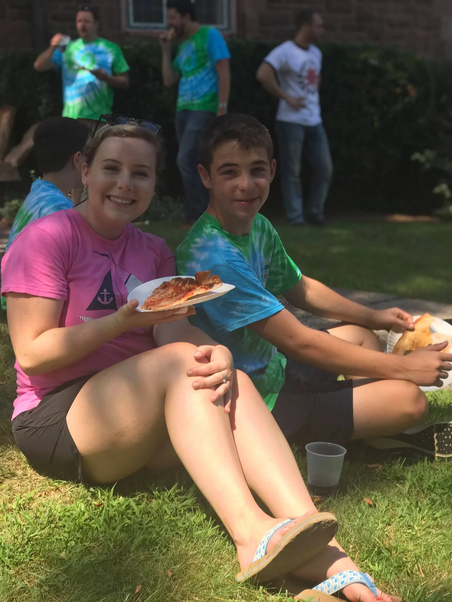 A group of young people having a picnic outside, sitting on grass, with two people dining on pizza in the foreground and others standing in the background.