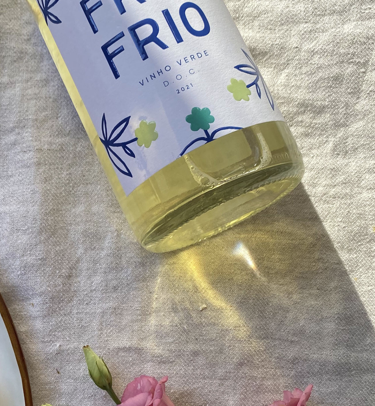 A bottle of white wine labeled 'Fii Fii RIO' resting on a light-colored fabric surface, casting a shadow, with pink flowers at the bottom edge of the image.